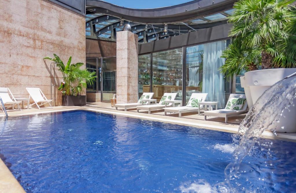 Se ve una piscina azul con una fuente y tumbonas blancas alrededor, bajo una estructura acristalada.