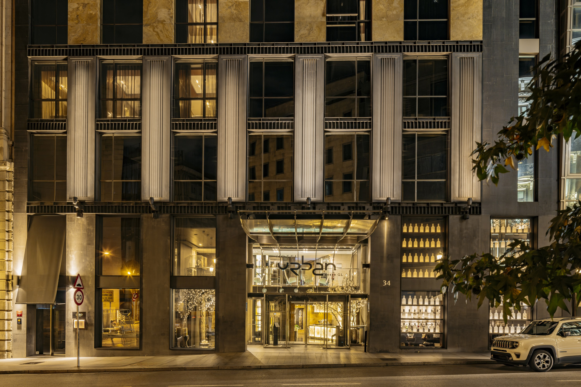 Se ve la fachada de un edificio con columnas y ventanas iluminadas, junto a la entrada principal y un vehículo estacionado en la calle.