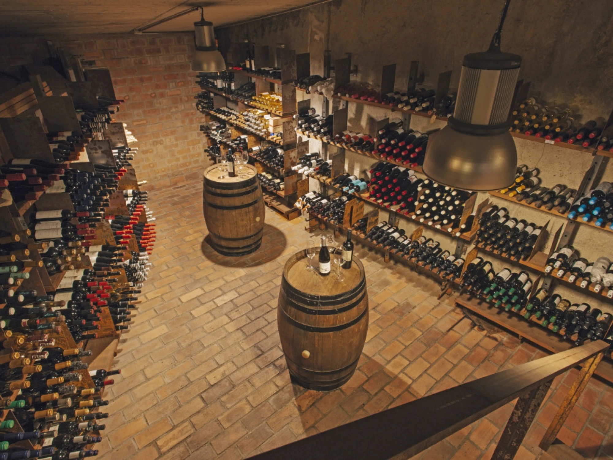 Se ve una bodega con barricas de madera y estanterías llenas de botellas, iluminadas con lámparas colgantes.