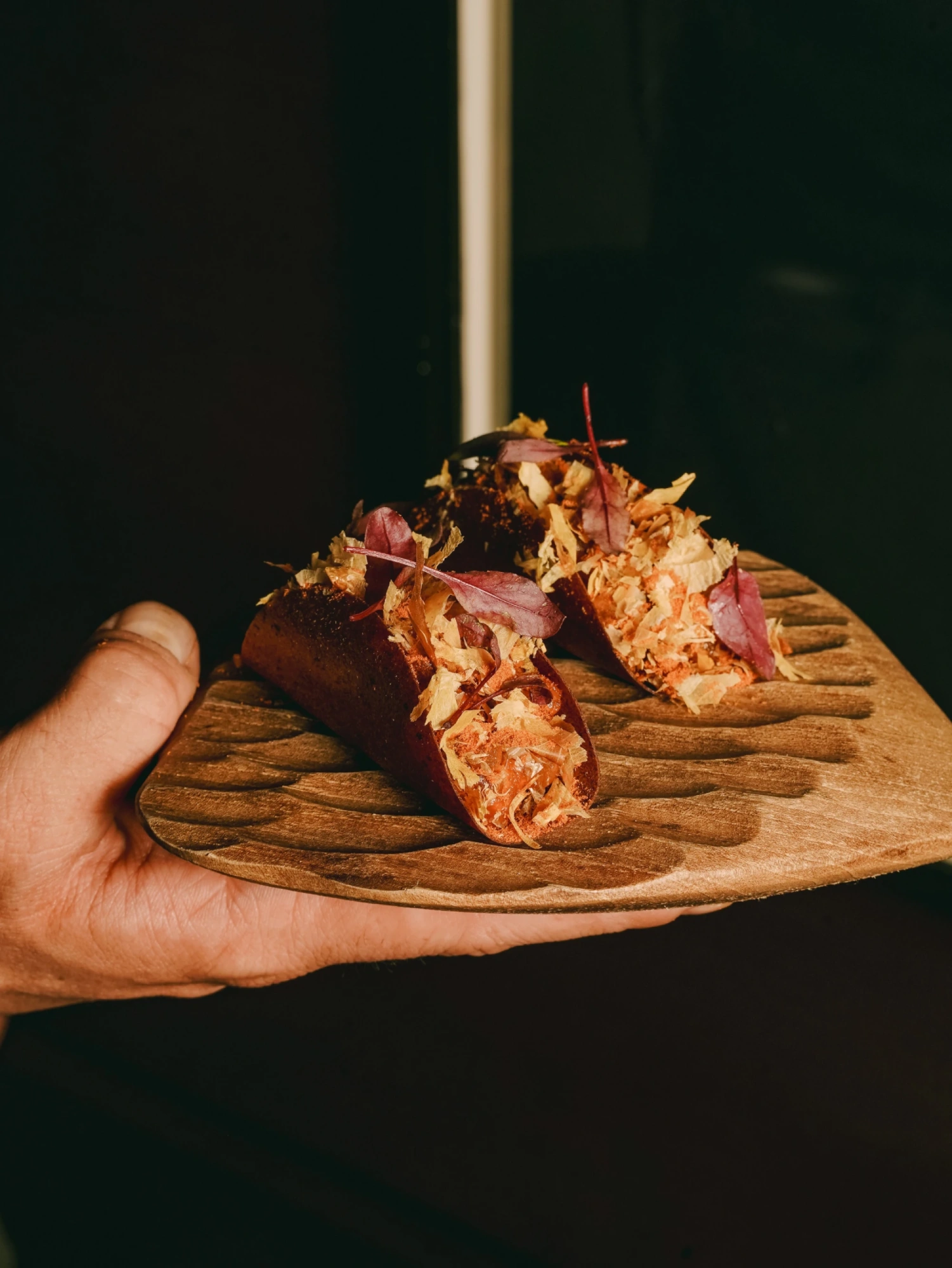 Una mano sostiene un plato con una tapa de comida decorada con atún rojo sobre una base de taco de maíz.
