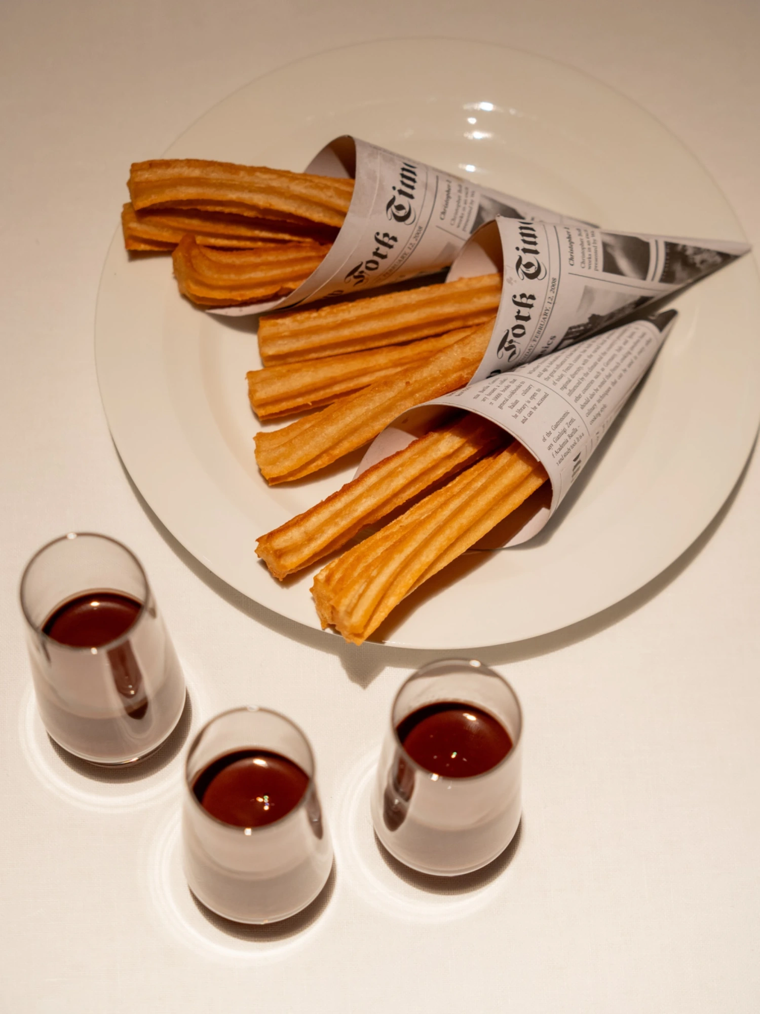 Se ve un plato con churros y tres vasos pequeños con chocolate sobre una mesa blanca.