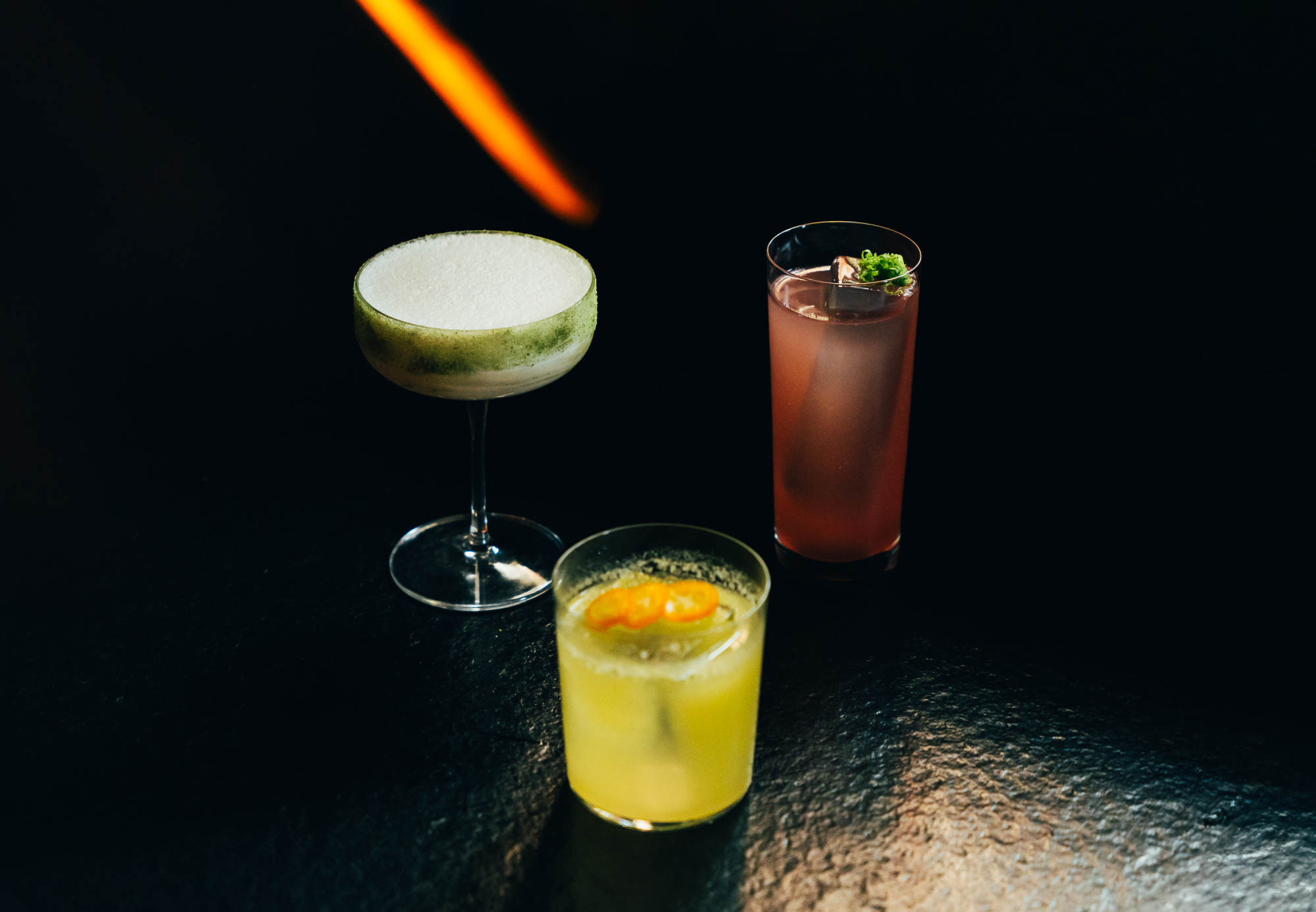 Se ven tres cócteles en vasos sobre una superficie oscura, con una guarnición verde en uno de ellos.
