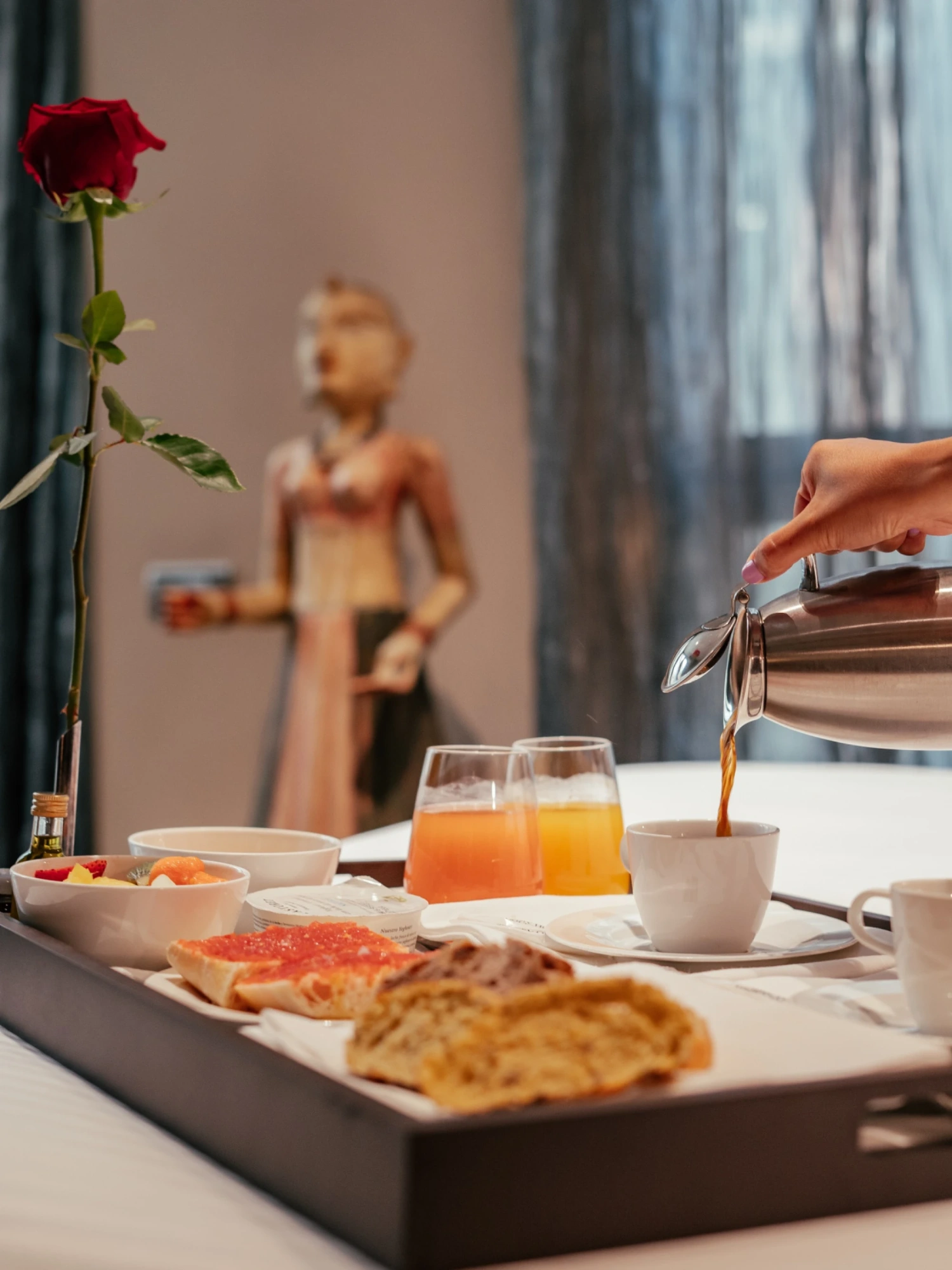 Se ve una bandeja con zumo en vaso, café, pan y un plato de comida junto a una figura decorativa en una habitación.