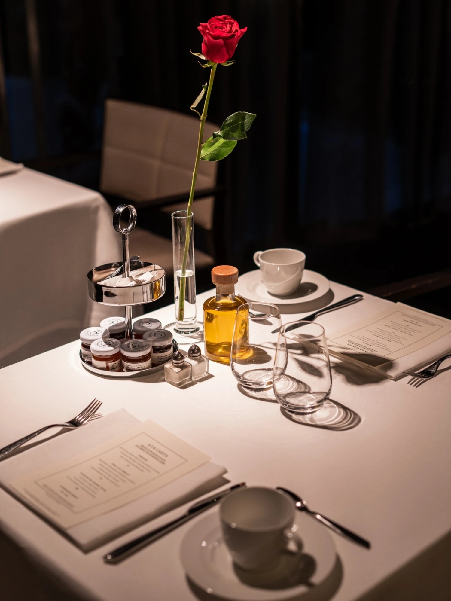 Se ve una mesa puesta con vajilla, cubiertos, una rosa en un florero y una botella con bebida.