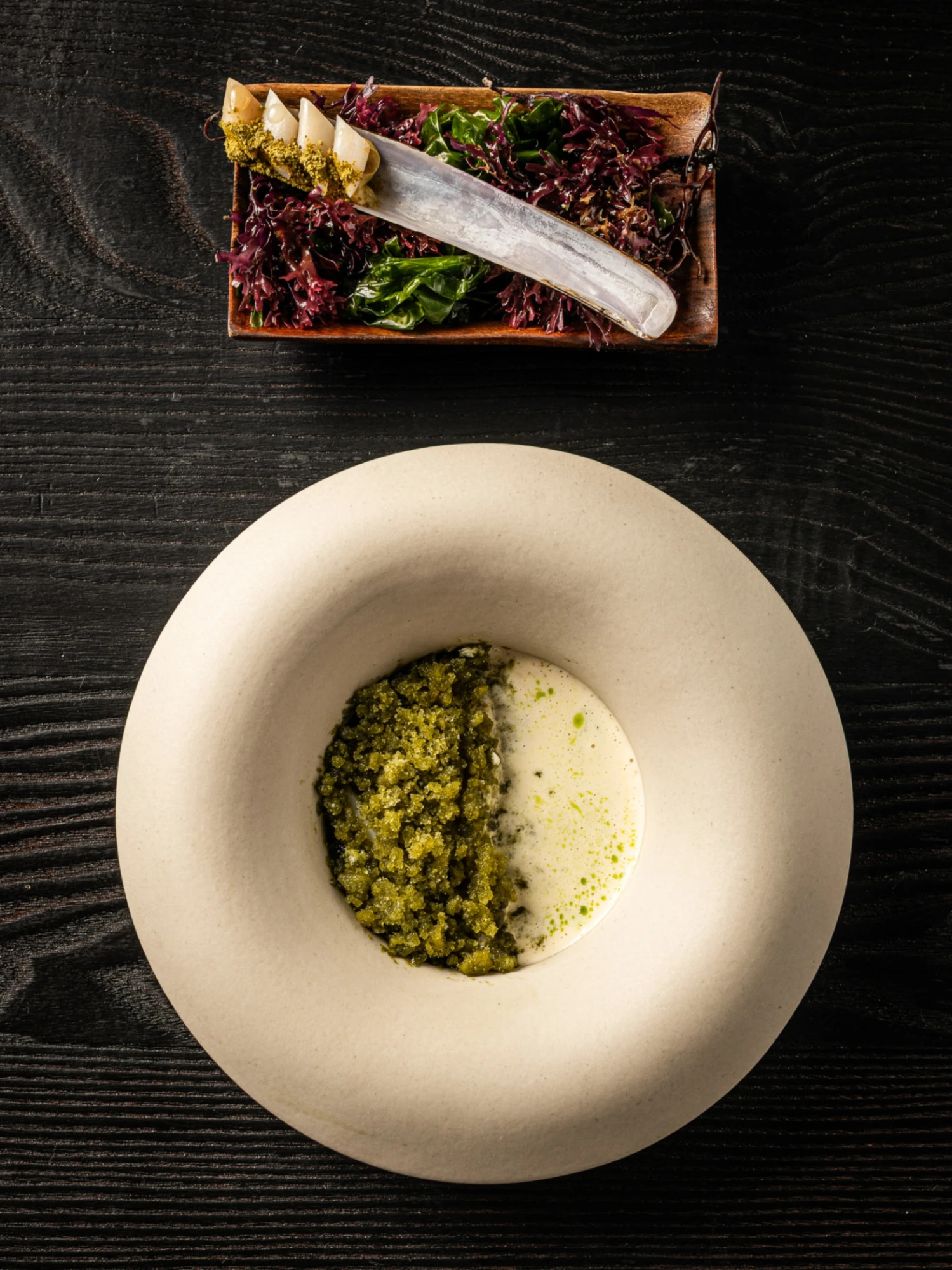 Un plato blanco con una porción de comida verde y un fondo oscuro con un emplatado adicional en la parte superior.