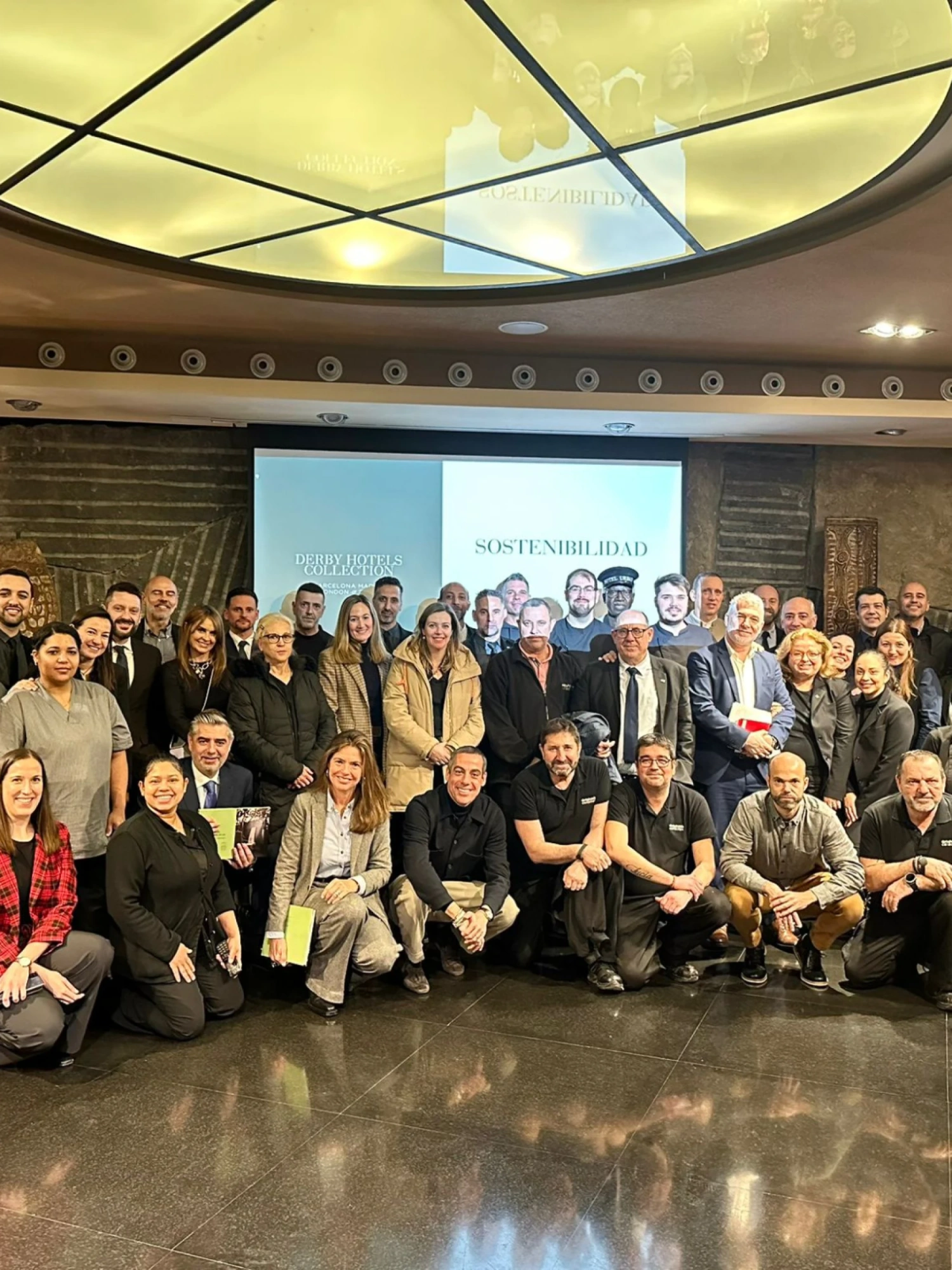 Un grupo de personas posa frente a una pantalla en una sala con suelo pulido y techo con iluminación.