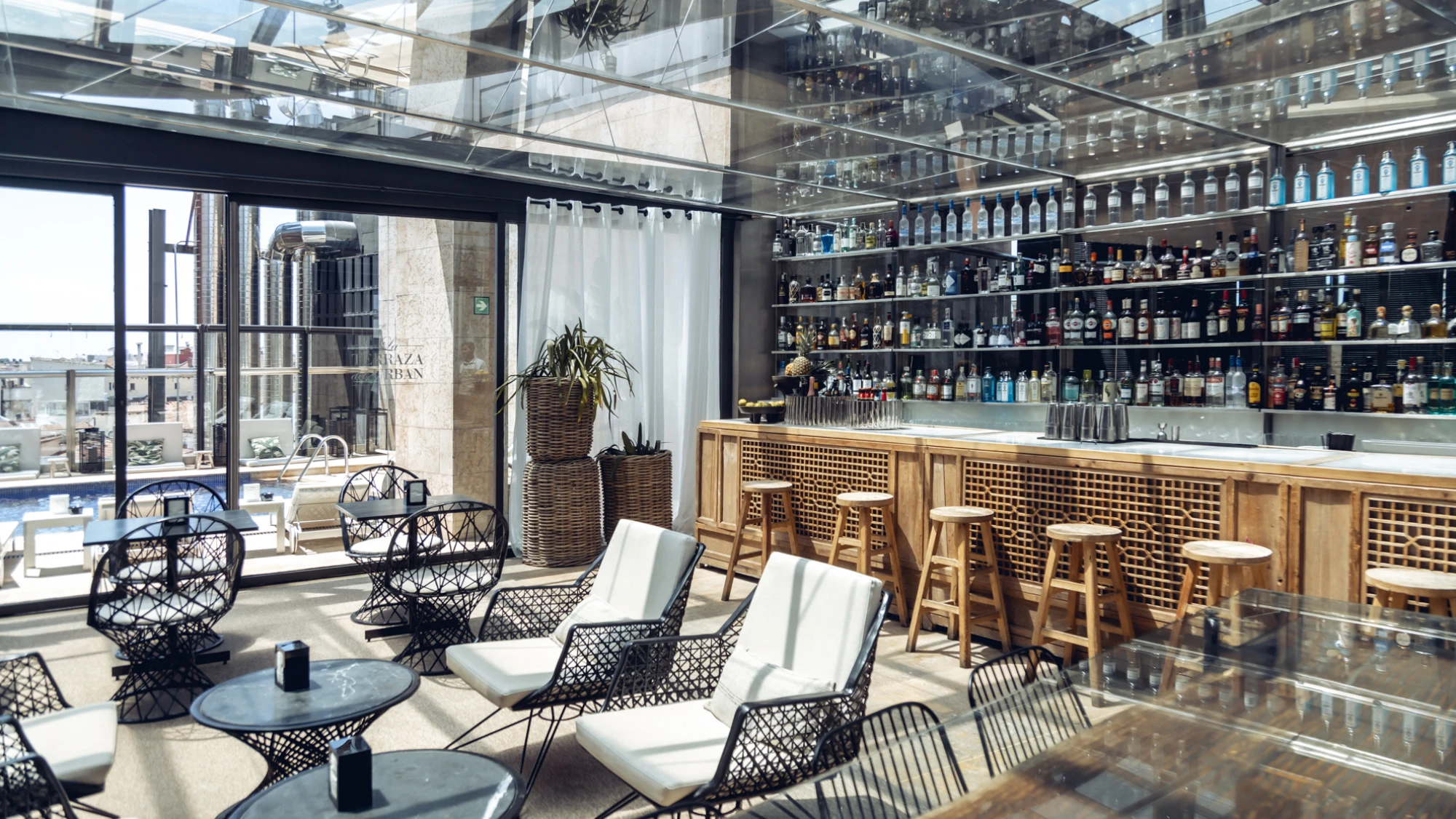 Se ve una terraza con mesas, sillas y una barra con botellas detrás, junto a barandillas de vidrio.