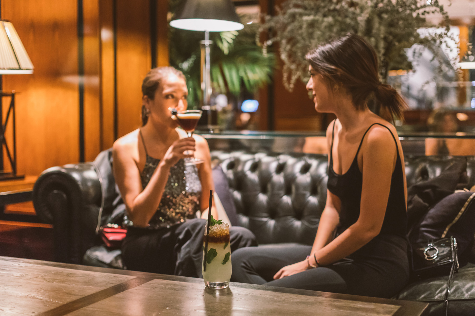 Two people are chatting while seated in a bar with drinks and a vase on the table.