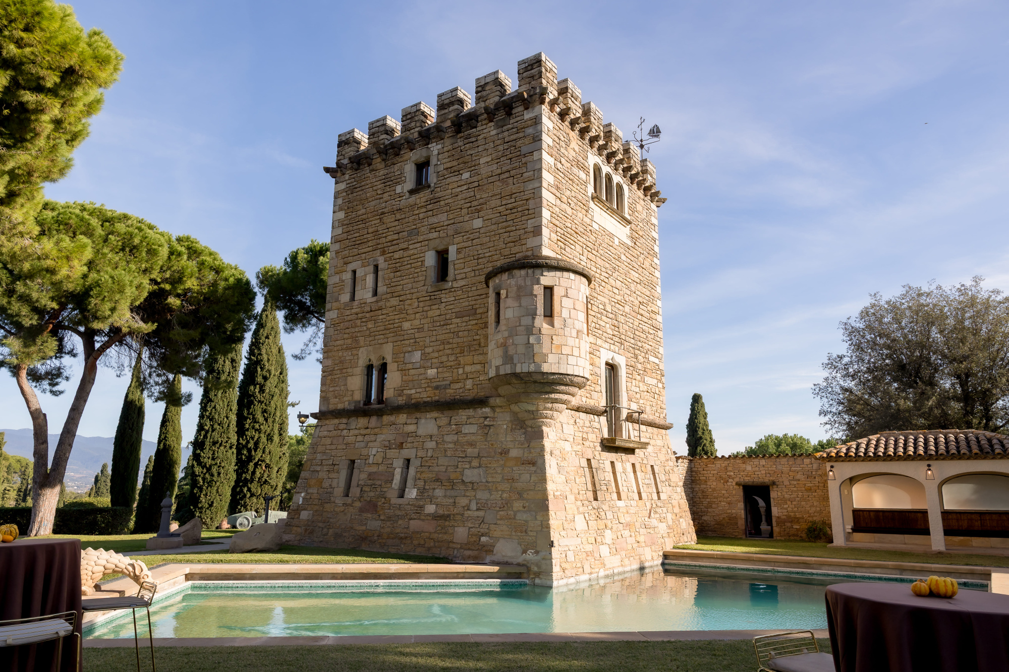 Se ve una torre de piedra junto a una piscina al aire libre y un jardín con árboles.
