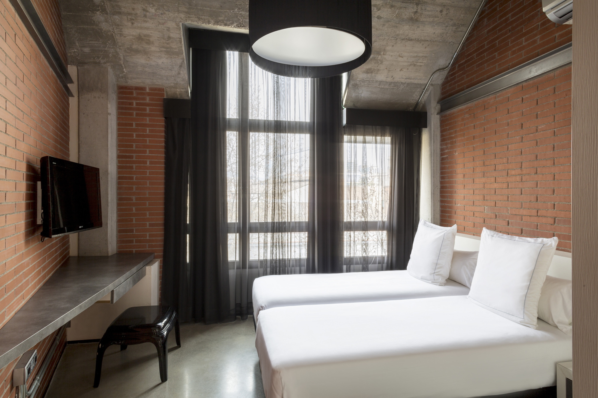Dormitorio con cama doble, almohadas blancas y grandes ventanas con cortinas oscuras.