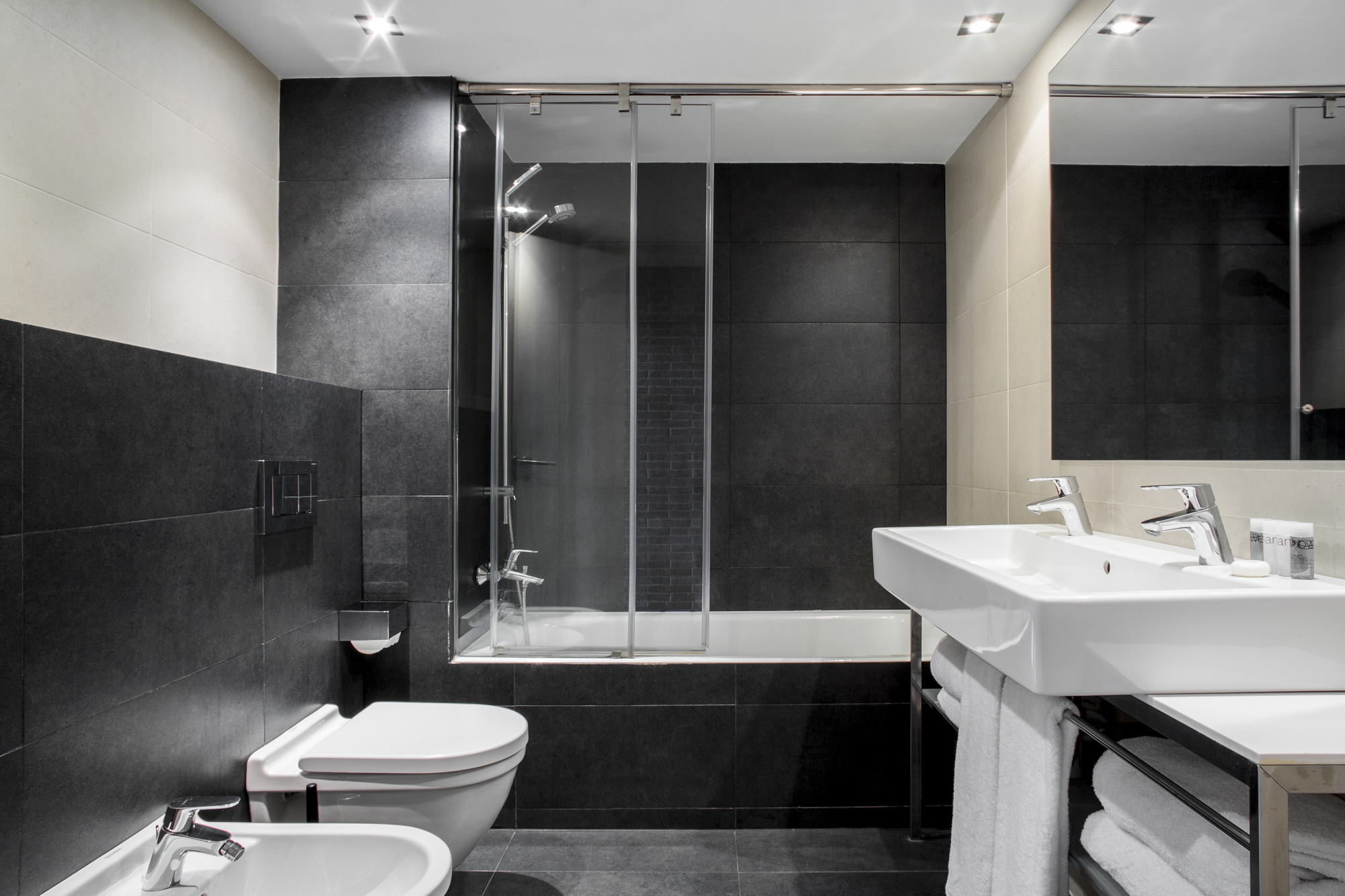 Baño moderno con ducha de cristal, inodoro y lavabo doble sobre encimera blanca.