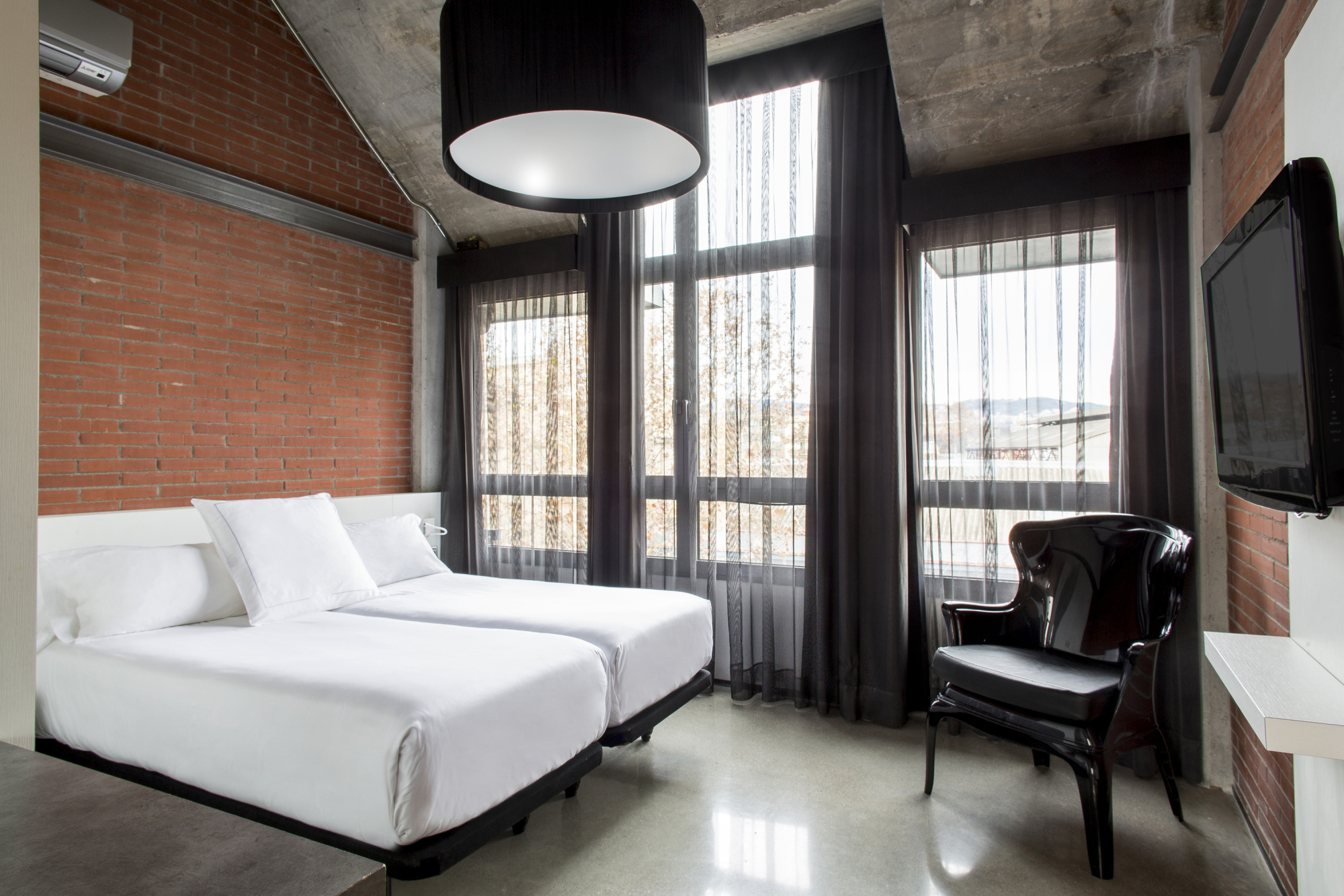 Se ve una cama con ropa de cama blanca, una silla oscura y ventanas con cortinas en una habitación con suelo blanco y pared de ladrillo.