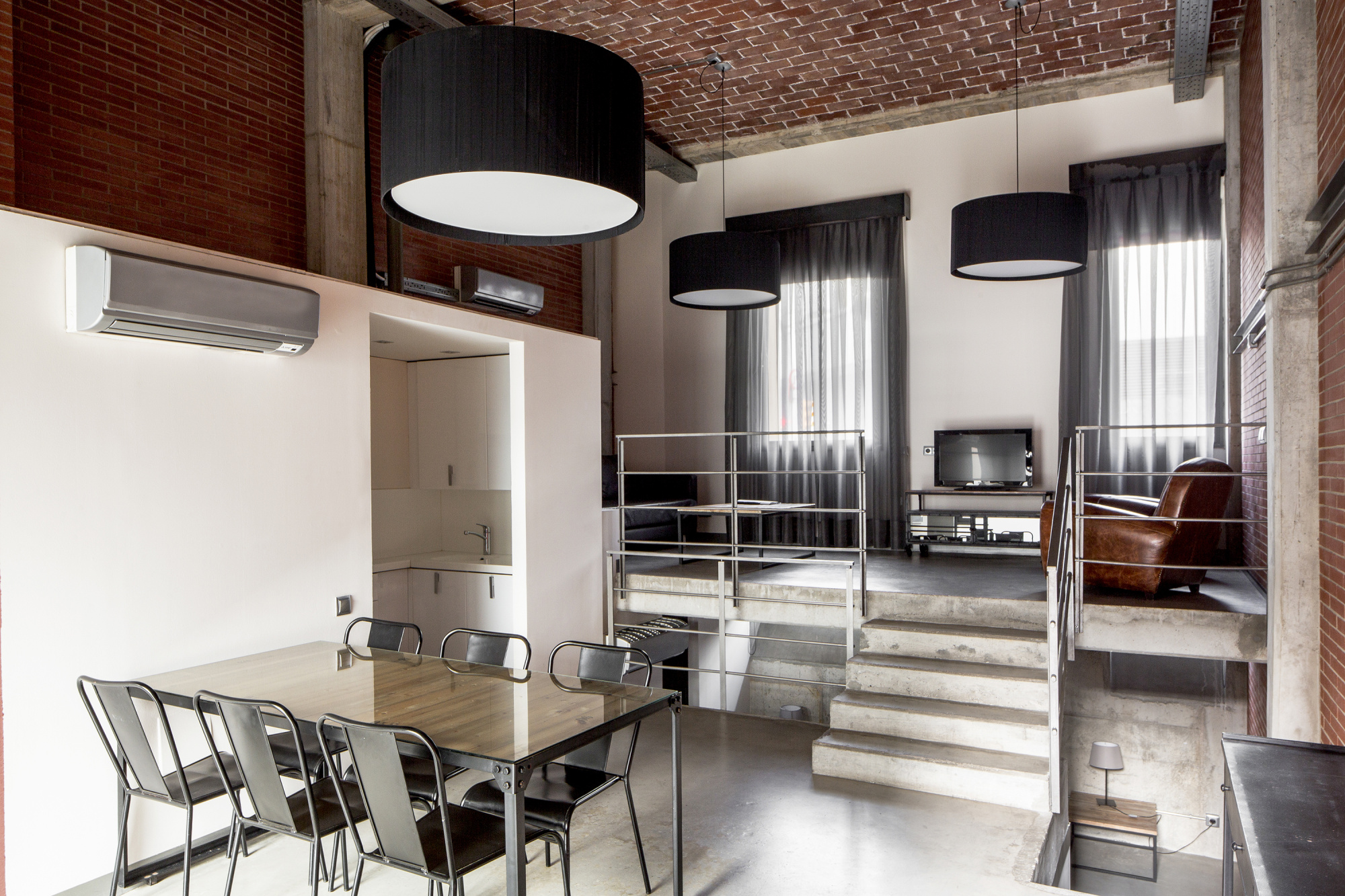 Se observa una zona de comedor con sillas metálicas y una cocina con encimera, junto a una sala con escaleras interiores.