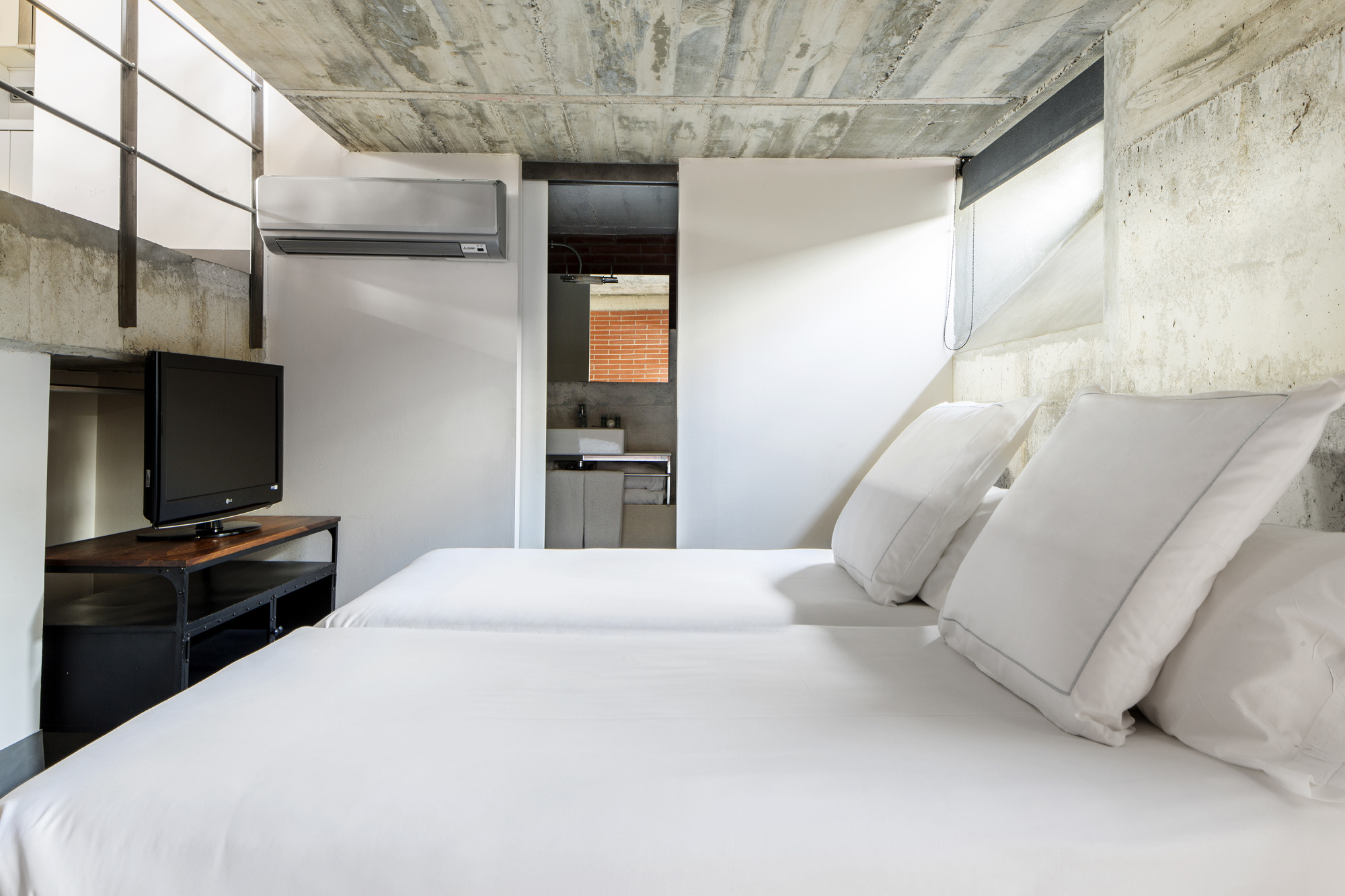 Se ve una cama doble con almohadas sobre ropa blanca y un televisor en un mueble, junto a una pared de color claro.
