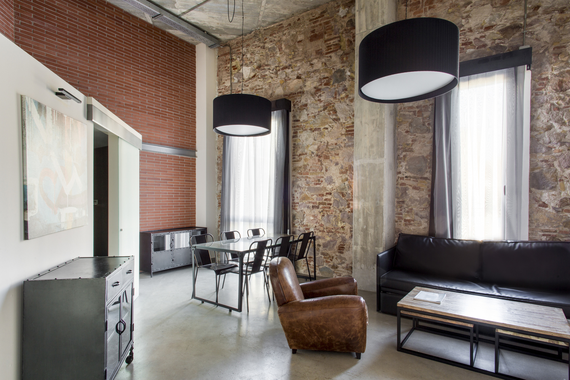 Se ve un salón con sofá marrón, mesa baja, lámparas de techo y paredes de ladrillo y piedra junto a ventanas con cortinas.