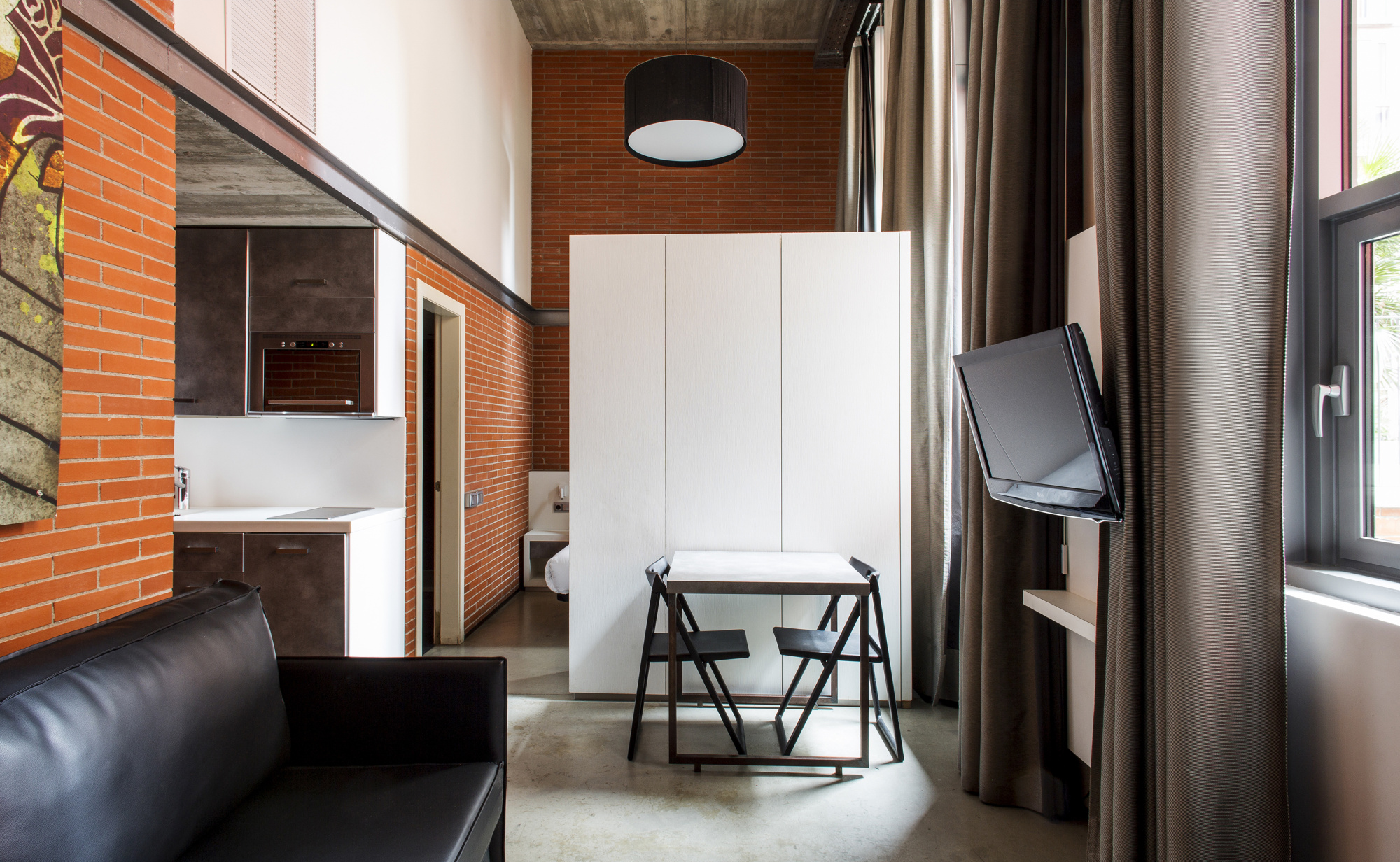 Se ve un estudio con sofá negro, escritorio y silla, armario blanco y una ventana con cortinas oscuras.