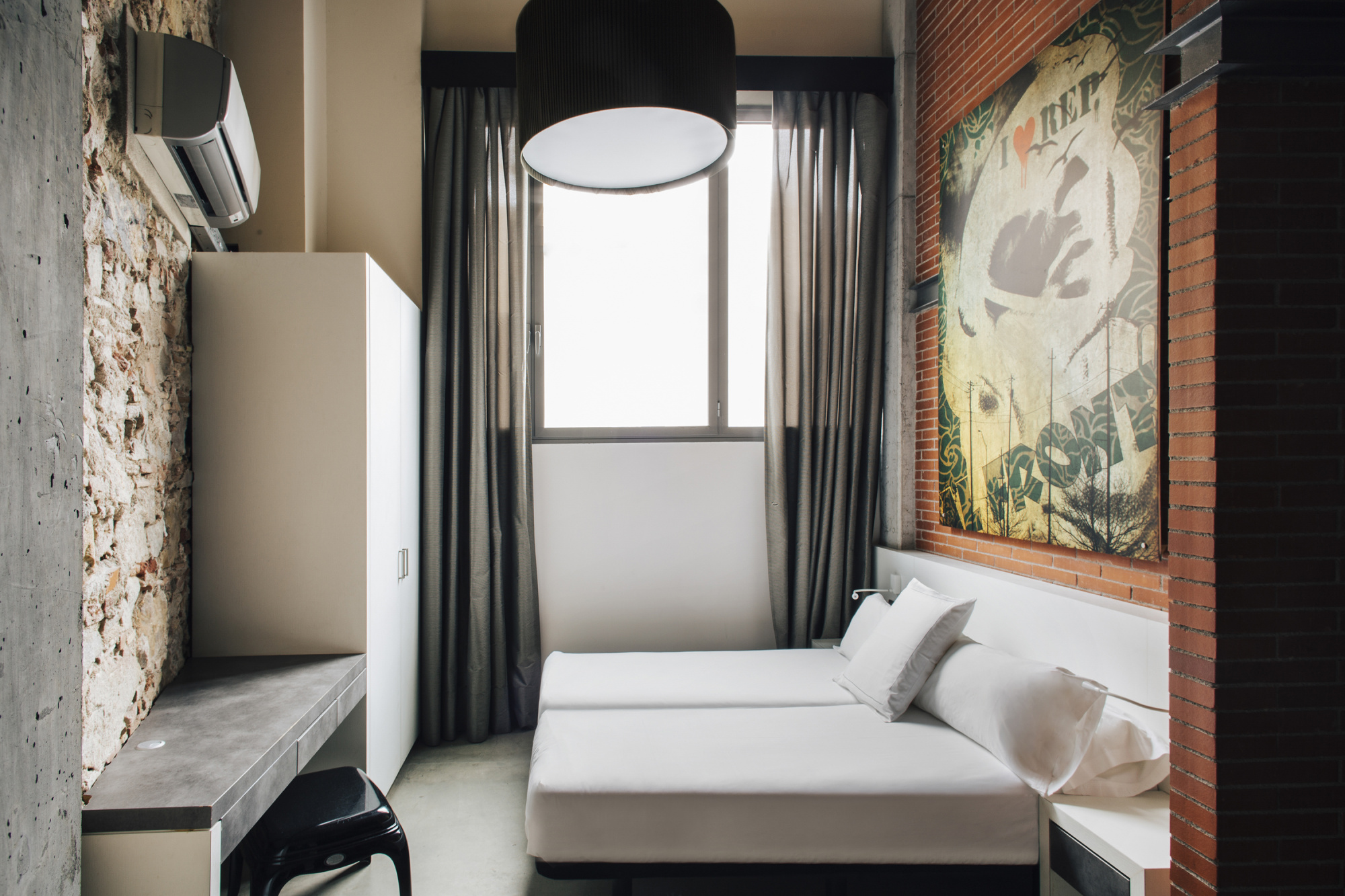 Se ve una cama individual con almohadas, una lámpara de techo y cortinas junto a una pared decorada.