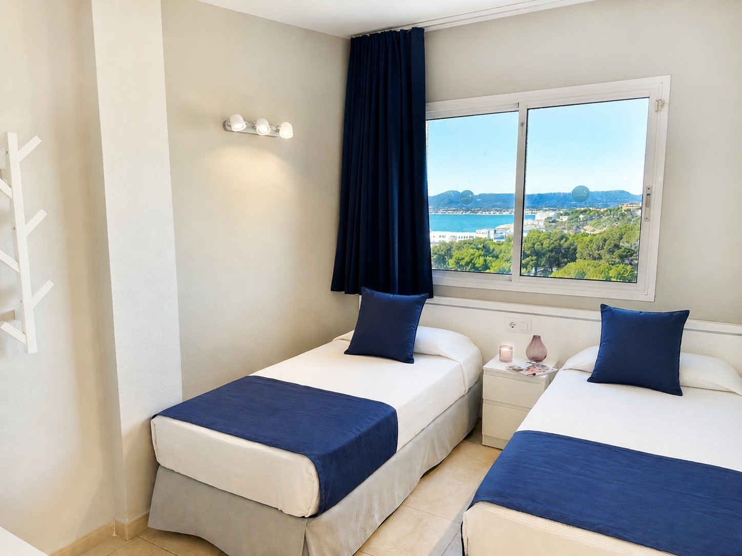 Se ve un dormitorio con dos camas individuales y almohadas azules junto a una ventana con cortinas oscuras.