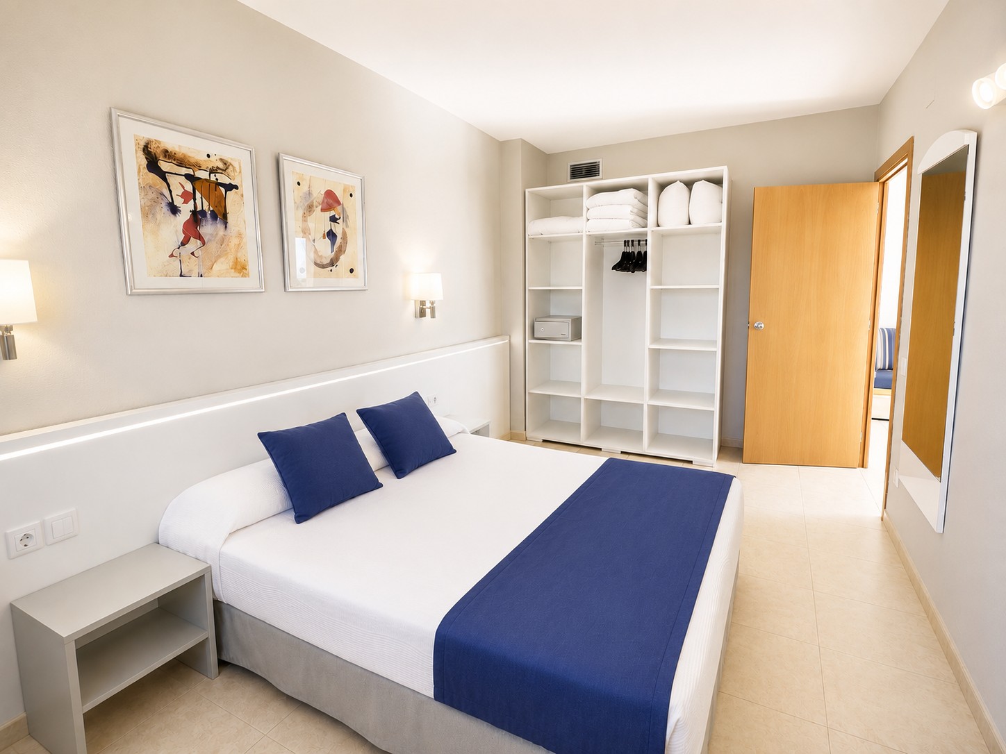 Se ve un dormitorio con una cama doble, mesita de noche, cuadros en la pared y un armario abierto con estantes.