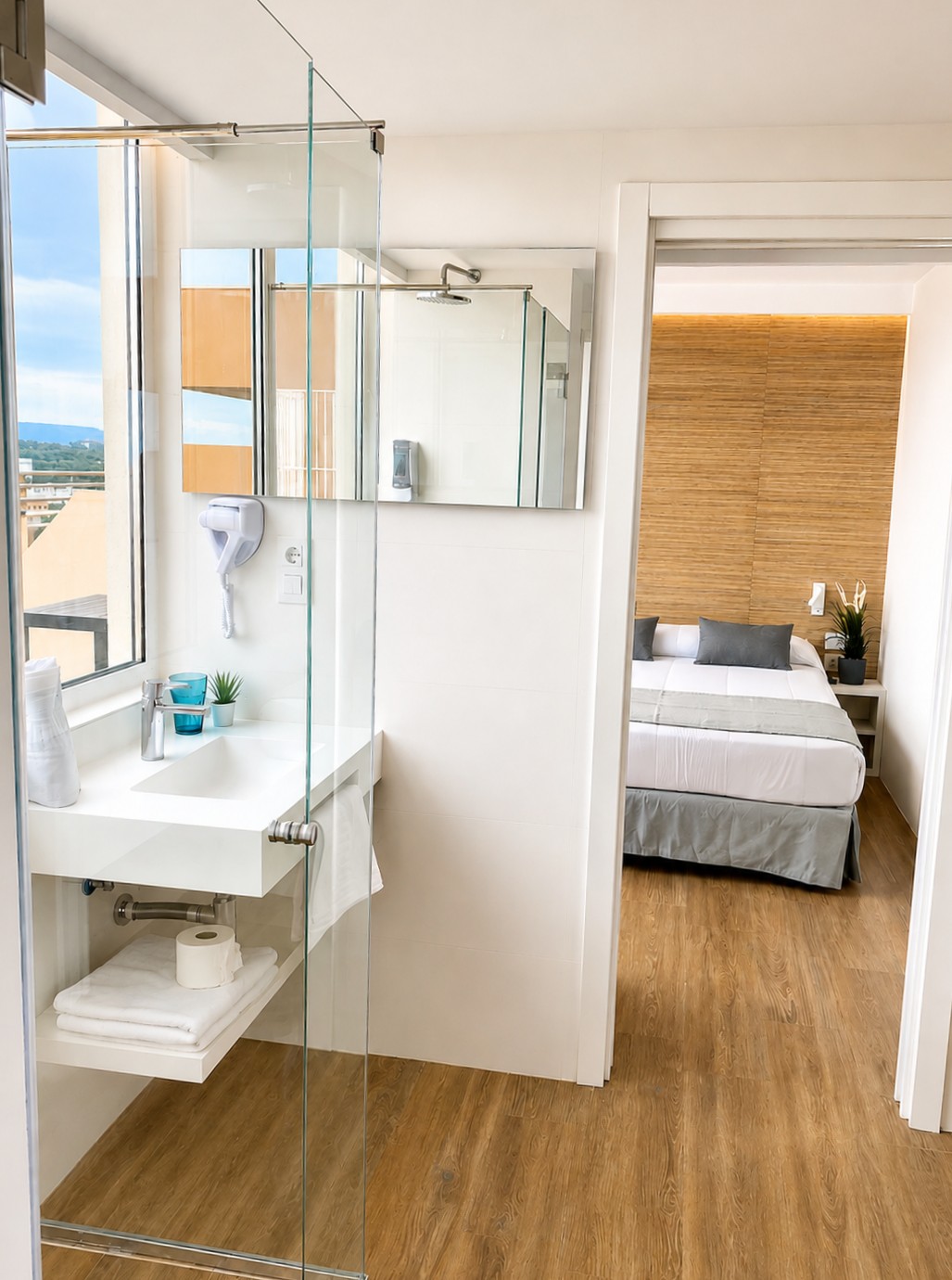 Se ve un baño con lavabo y ducha de cristal junto a una zona de dormitorio con cama y suelo de madera.
