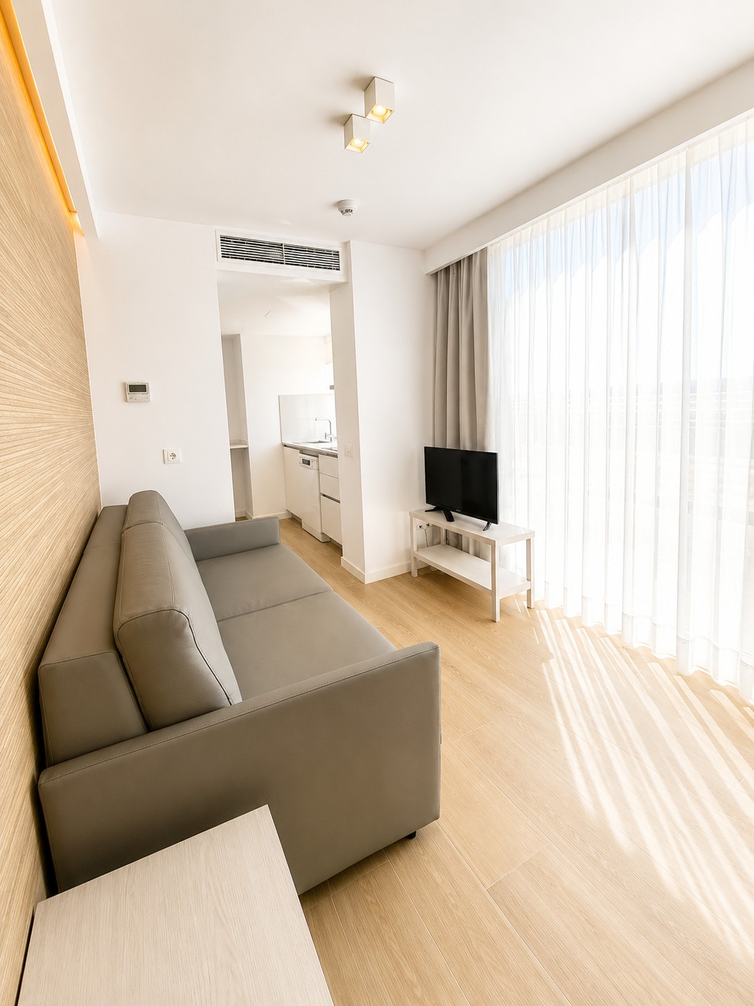 Se ve una sala luminosa con sofá, televisor y cortinas junto a una zona de cocina abierta.