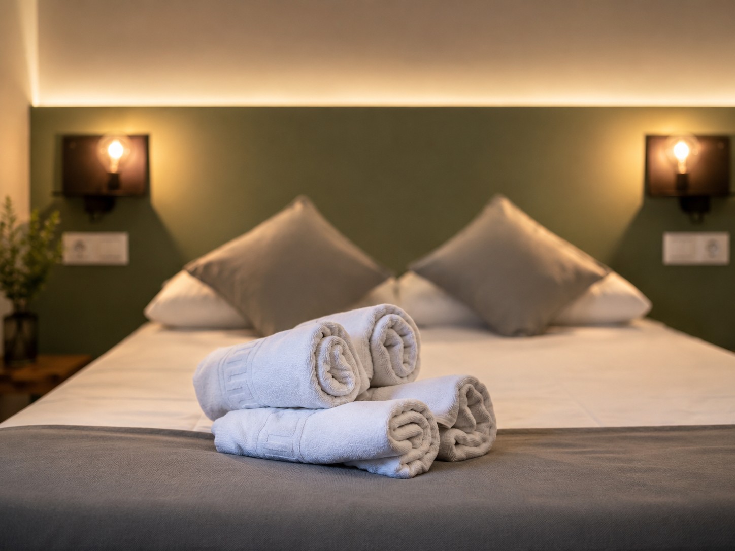 La imagen muestra una cama con almohadas y toallas dobladas sobre la colcha en una habitación con iluminación tenue.