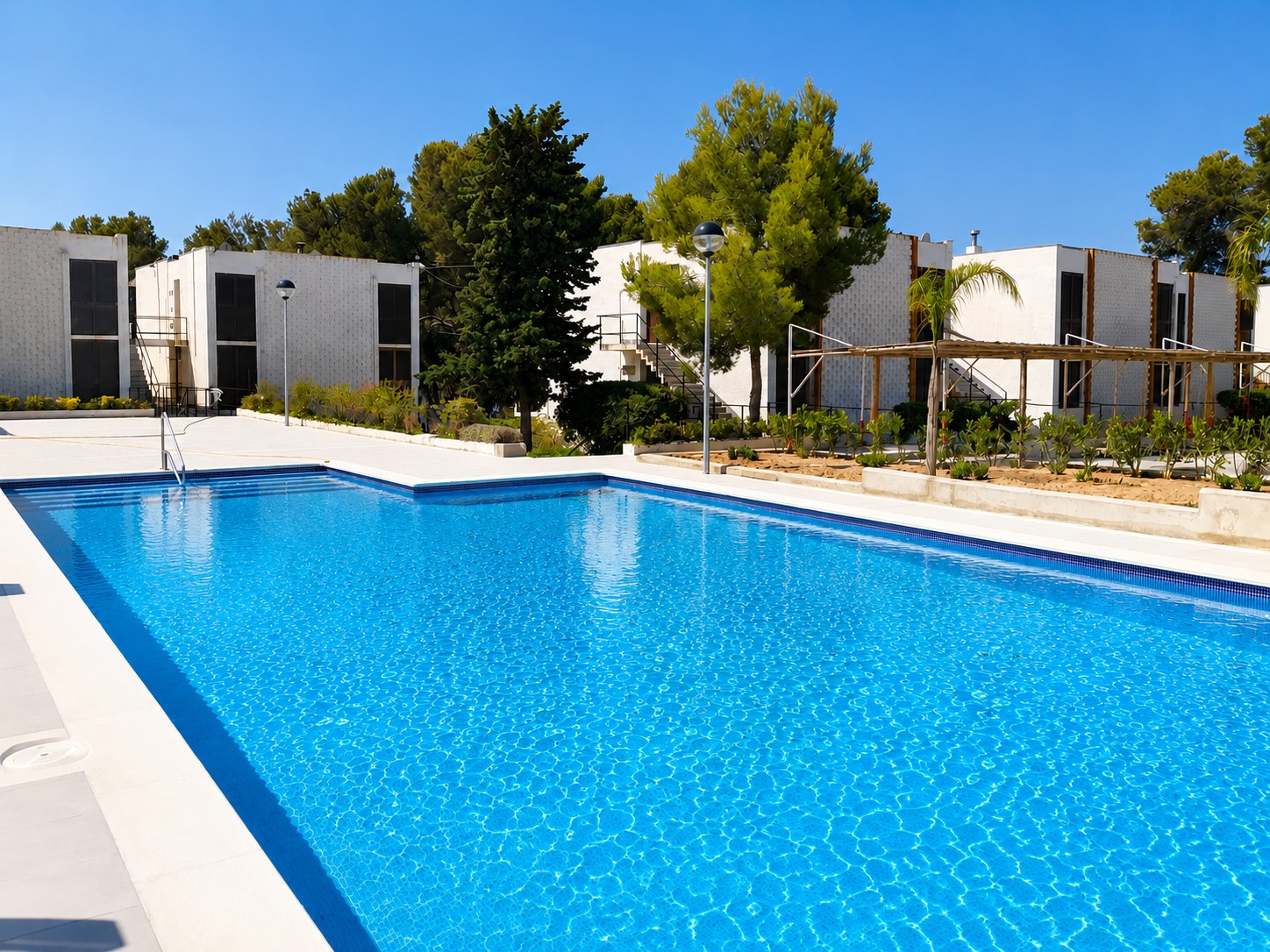 Se ve una piscina azul al aire libre junto a un área ajardinada y una zona de descanso con tumbonas.