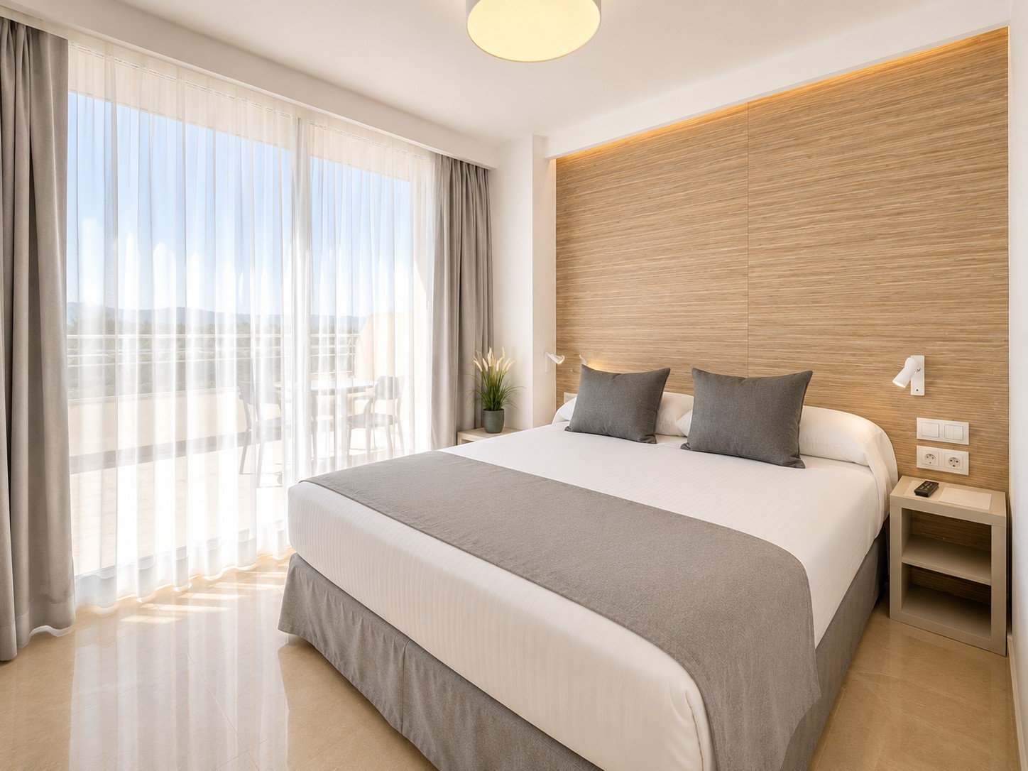 Se ve una cama doble con ropa de cama clara, dos almohadas y una mesita junto a una ventana con cortinas.