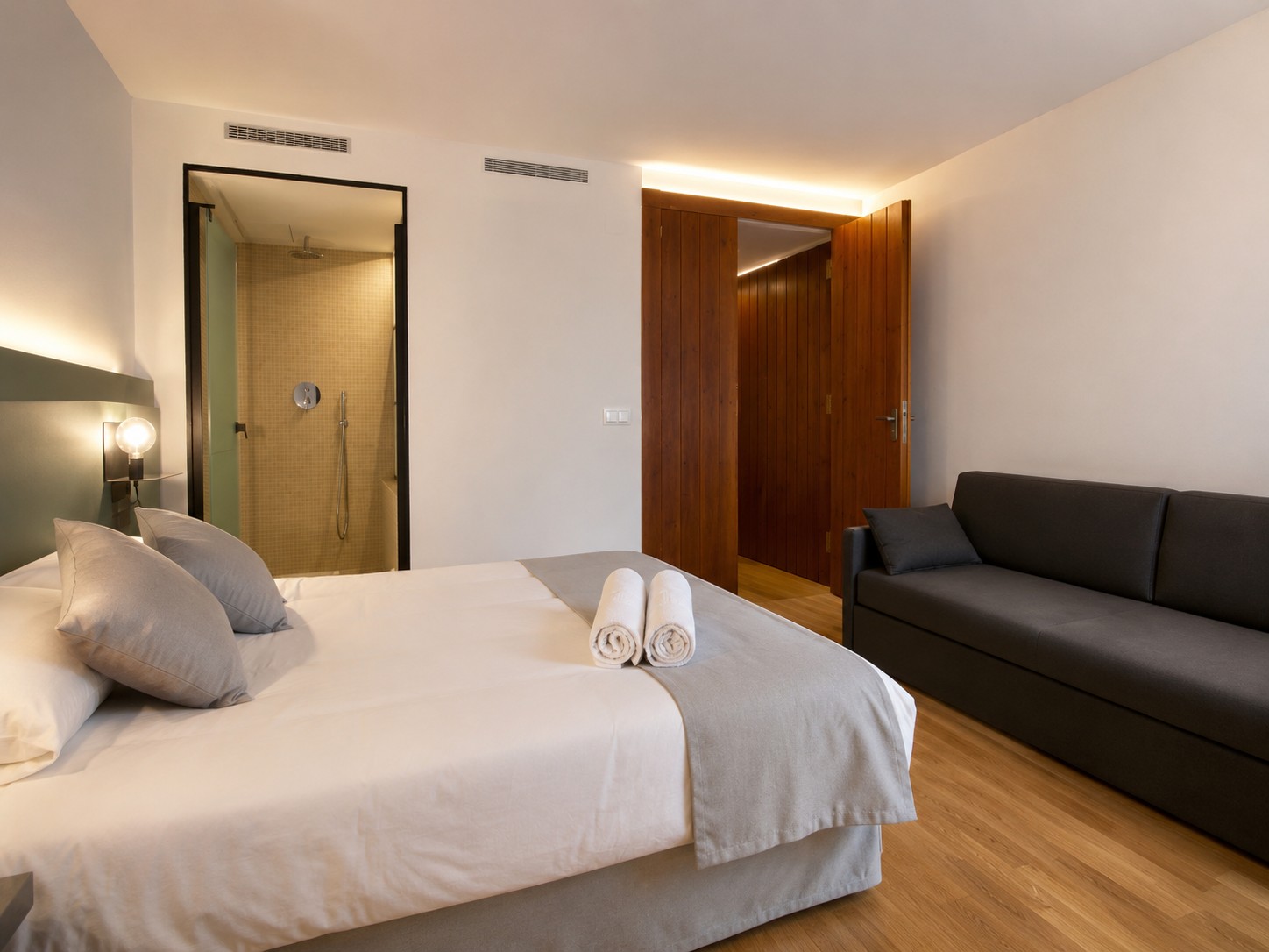 Se ve una cama con ropa de cama blanca, un sofá gris y una puerta de madera en el dormitorio.