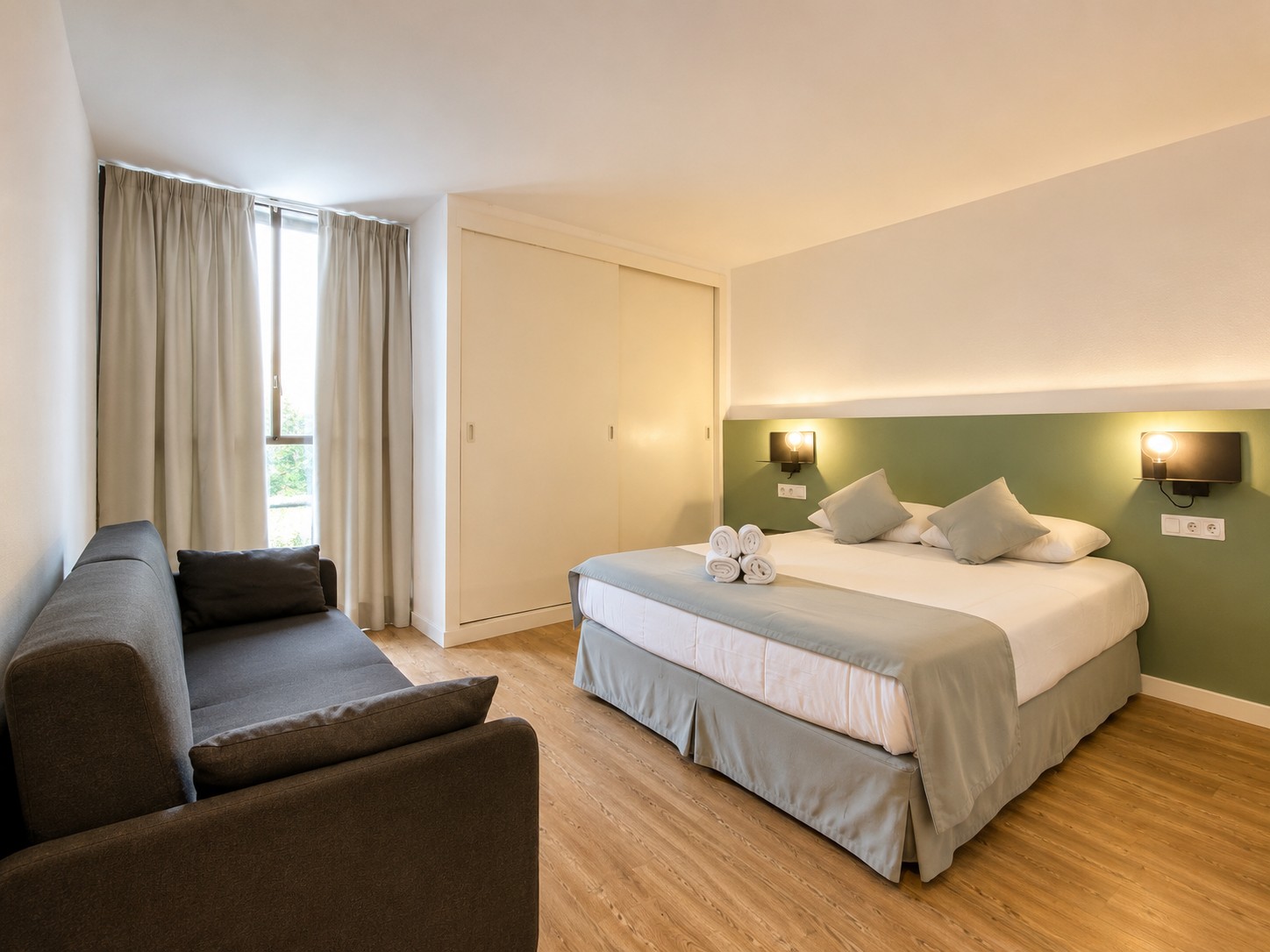 Se ve una cama doble con almohadas, una zona de estar con sofá y cortinas claras junto a una pared verde.