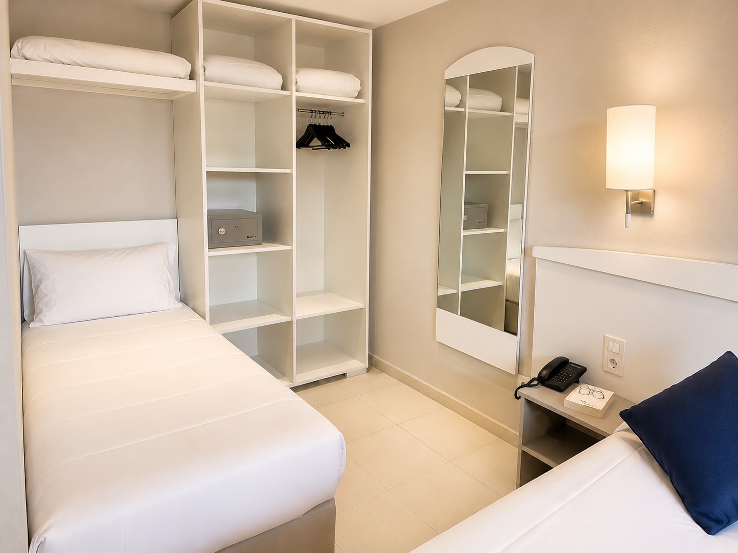 Se ve una cama individual con ropa de cama blanca y estanterías integradas, junto a una lámpara de pared y un escritorio.
