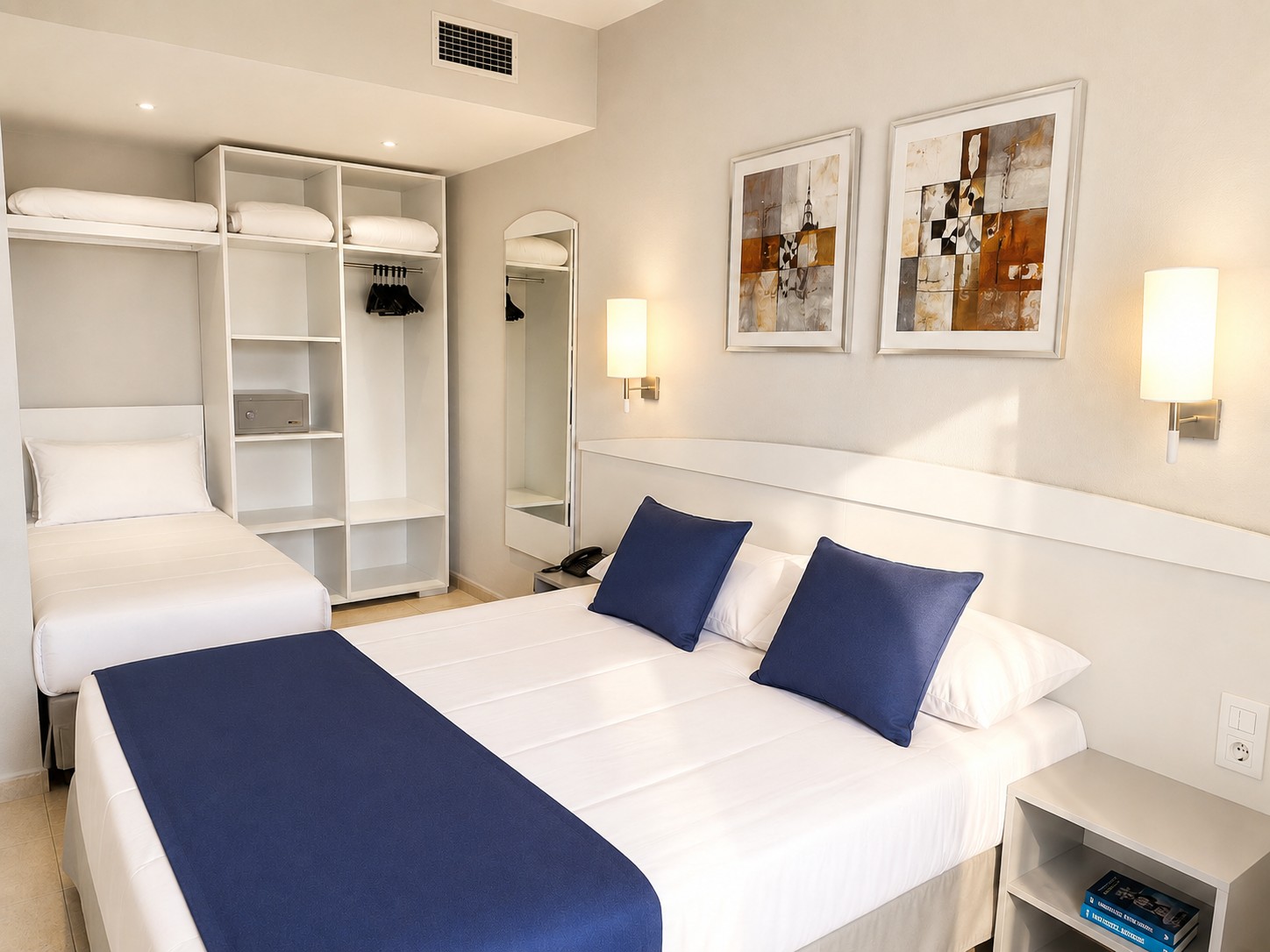 Se ve una cama doble con cojines azules, estanterías empotradas y lámparas de pared en una habitación luminosa.