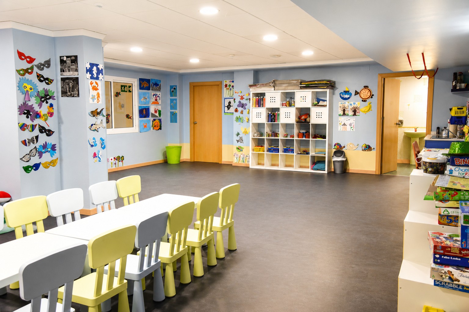 Sala infantil con mesas y sillas, estanterías y decoración mural en las paredes.