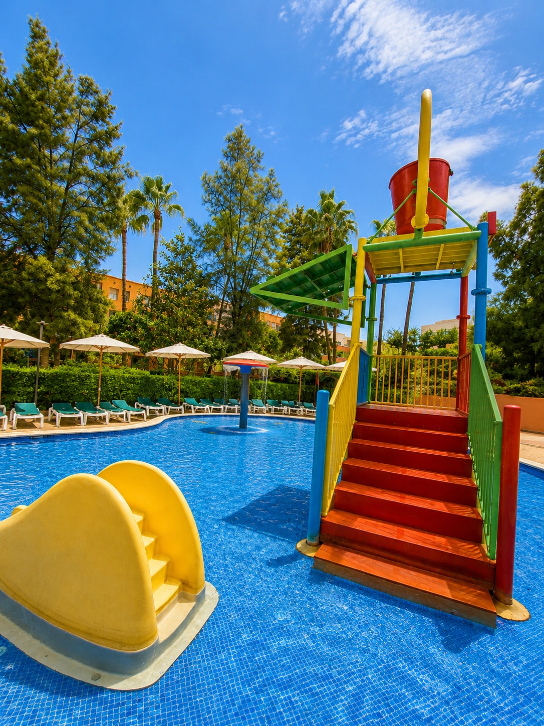 Se ve una piscina con zona infantil y un tobogán de colores junto a tumbonas y sombrillas.