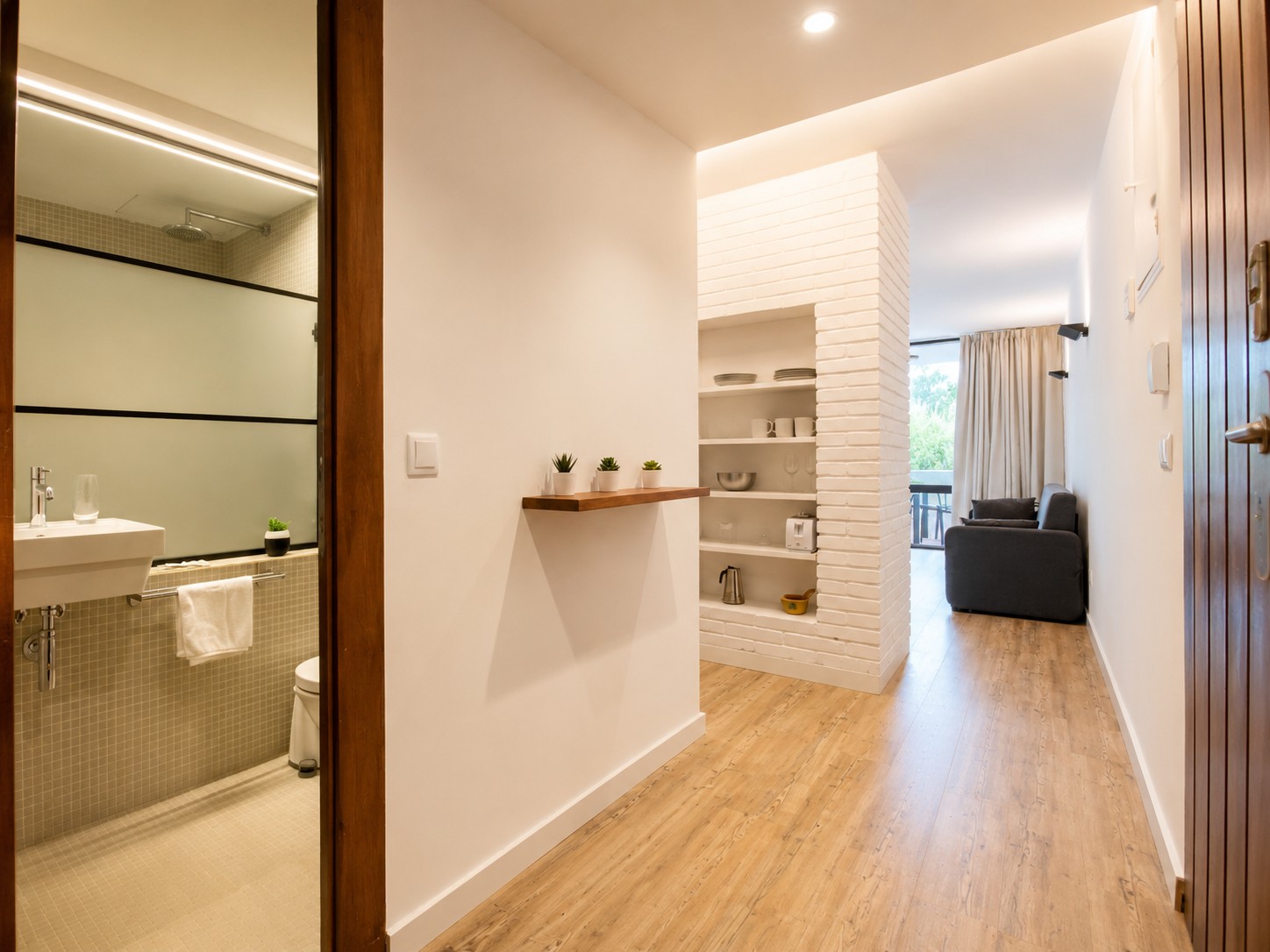 Se ve un pasillo con suelo de madera, paredes claras y una puerta abierta hacia una zona de estar con sofá.