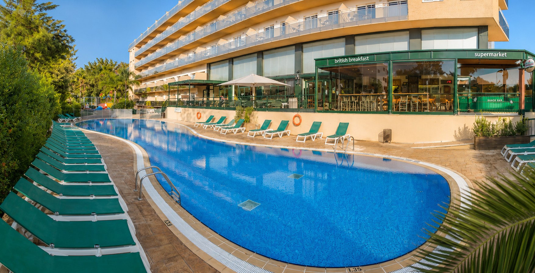 Se ve una piscina azul al aire libre junto a un edificio con terraza y sombrillas verdes.