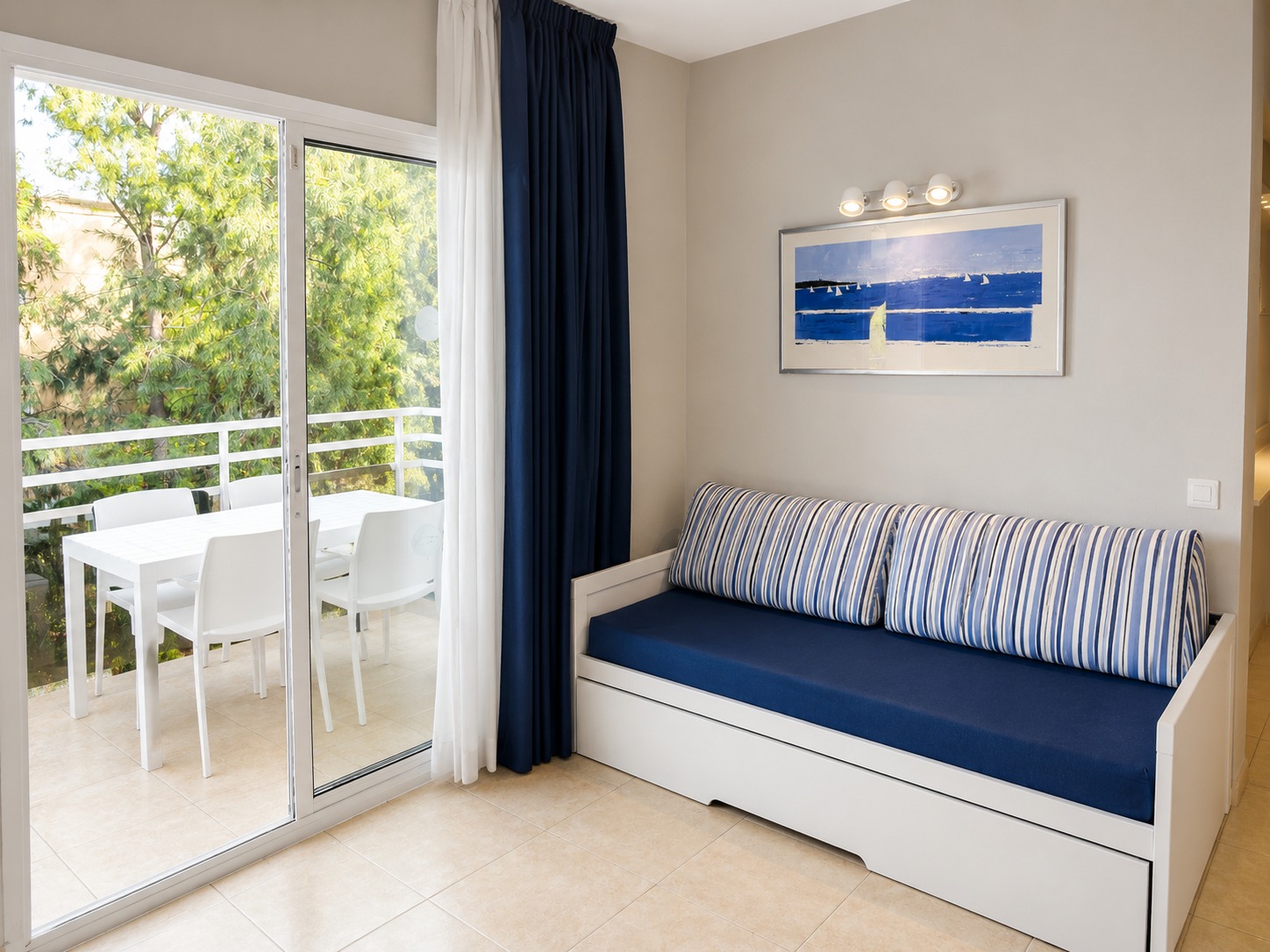Se ve un sofá cama junto a un ventanal con cortinas oscuras y una mesa blanca en un espacio luminoso.