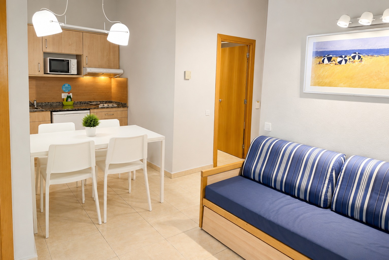 Se ve un sofá azul con cojines a rayas y una mesa junto a una zona de estar con iluminación de techo.