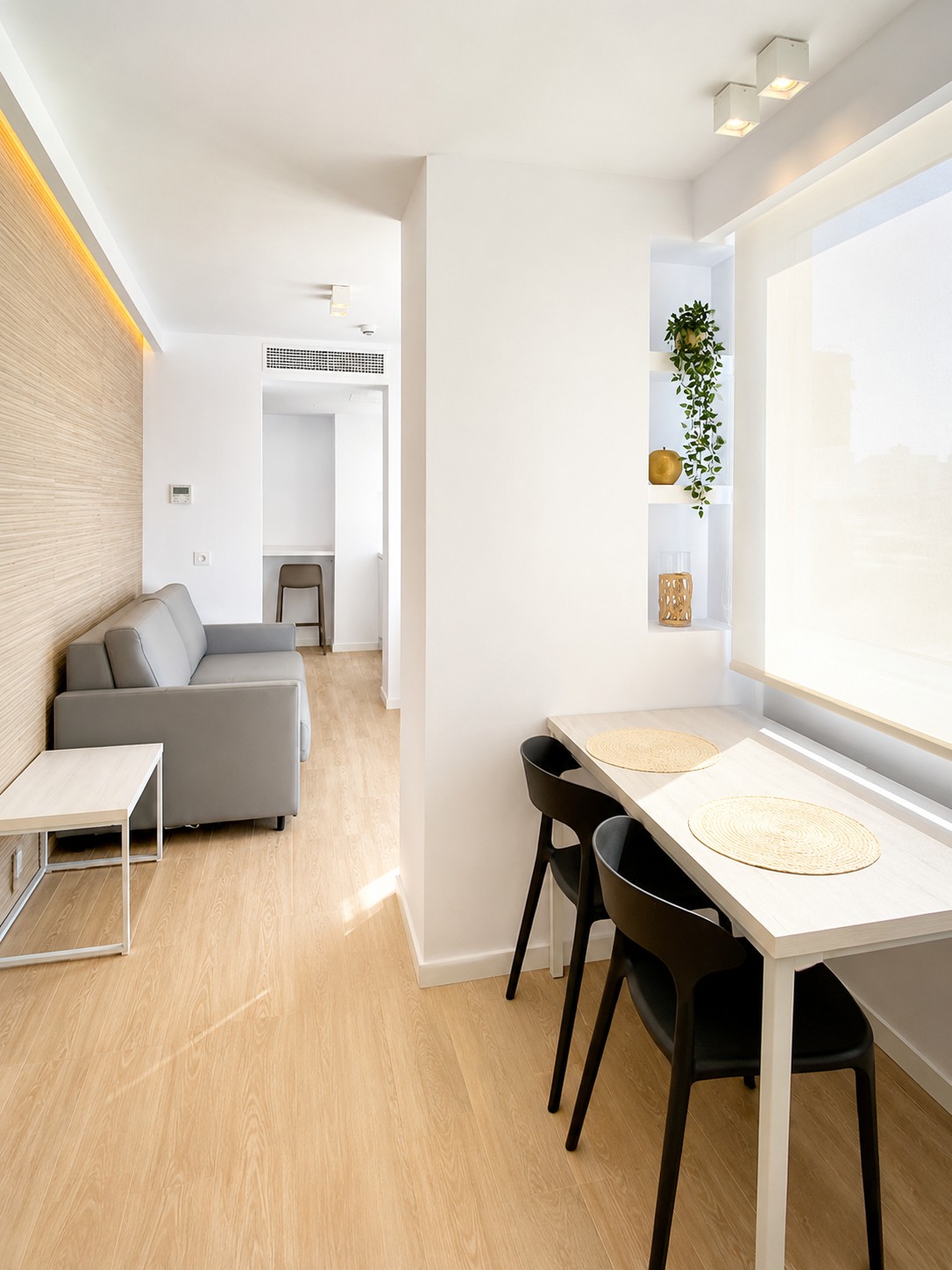 Se ve un salón con un sofá gris, una mesa auxiliar y una mesa de comedor con sillas junto a una pared blanca.