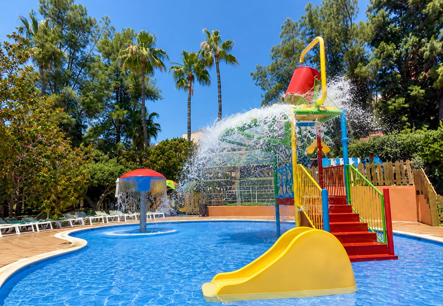Se ve una piscina al aire libre con una zona de juegos infantil y tobogán junto a vegetación.