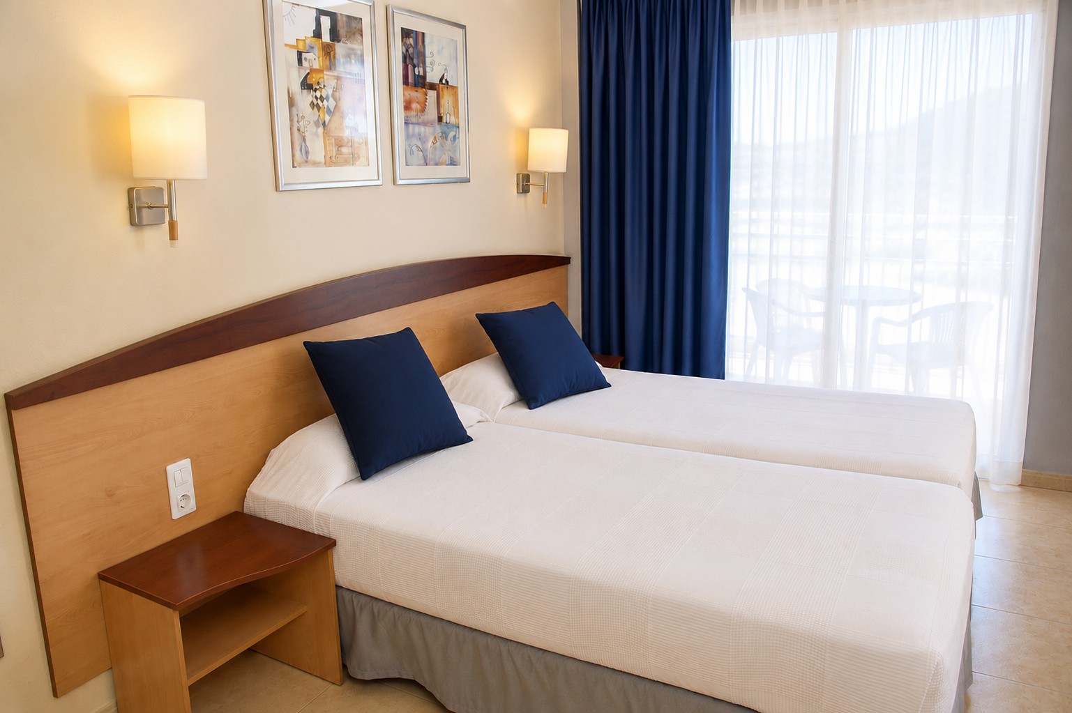 Se ve una cama doble con mesita y lámparas junto a una ventana con cortinas azules.