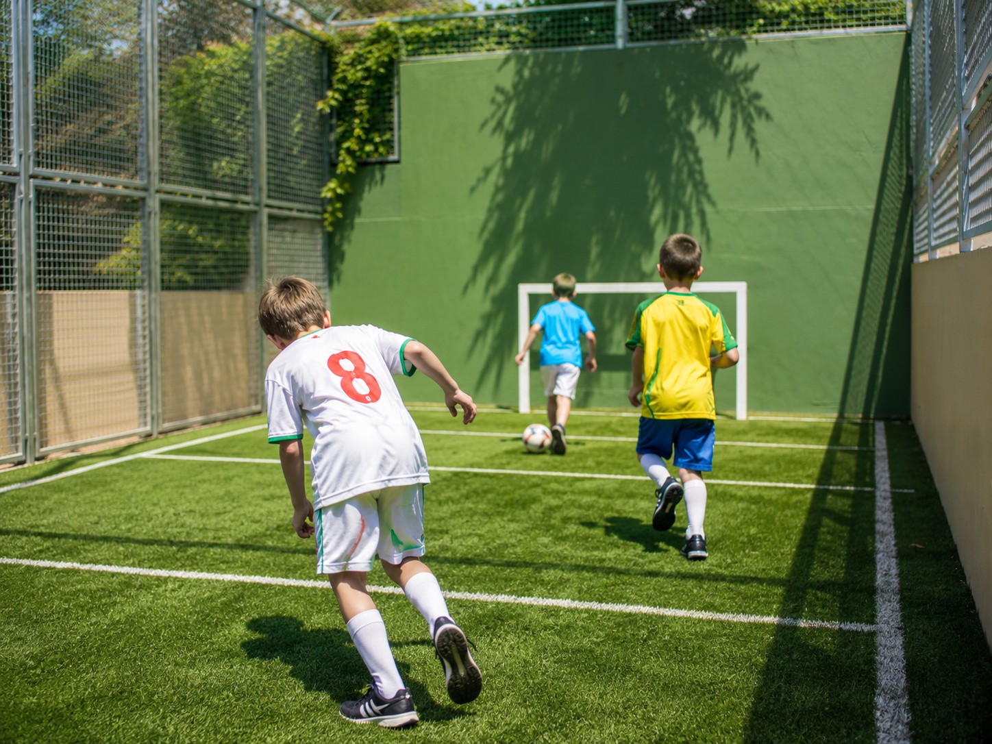 Niños juegan al fútbol en un campo exterior con césped artificial y porterías.