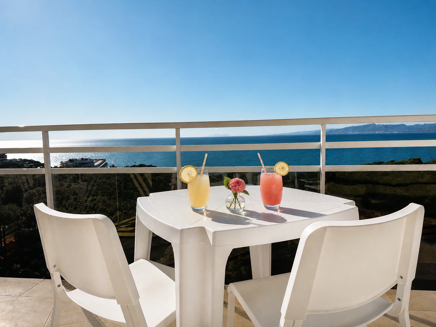 Se ve una terraza con una mesa blanca y cuatro sillas, con bebidas sobre la mesa y barandilla al fondo.