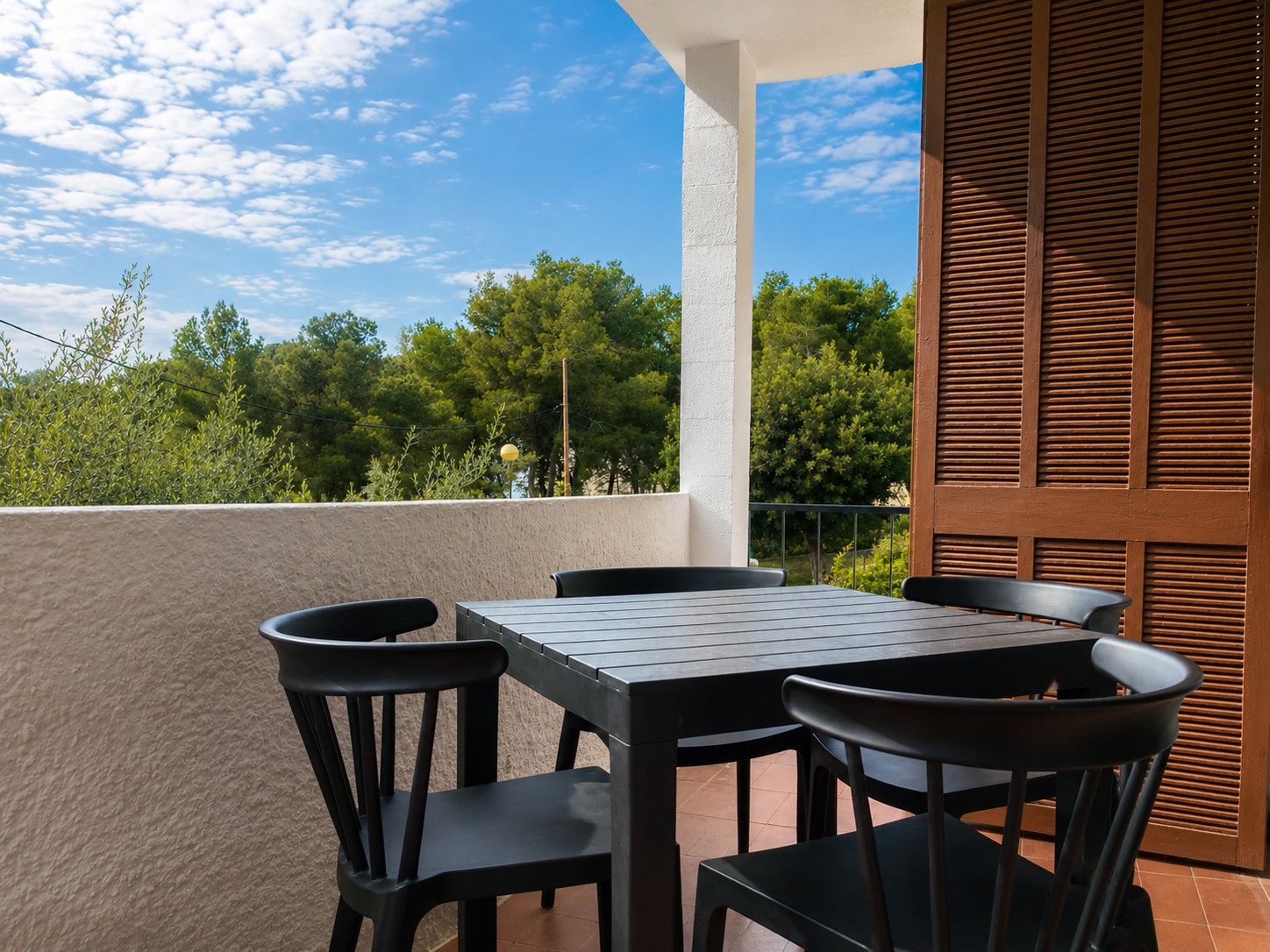 Se ve una terraza con mesa rectangular y sillas negras, junto a una pared y una puerta de madera con persianas.