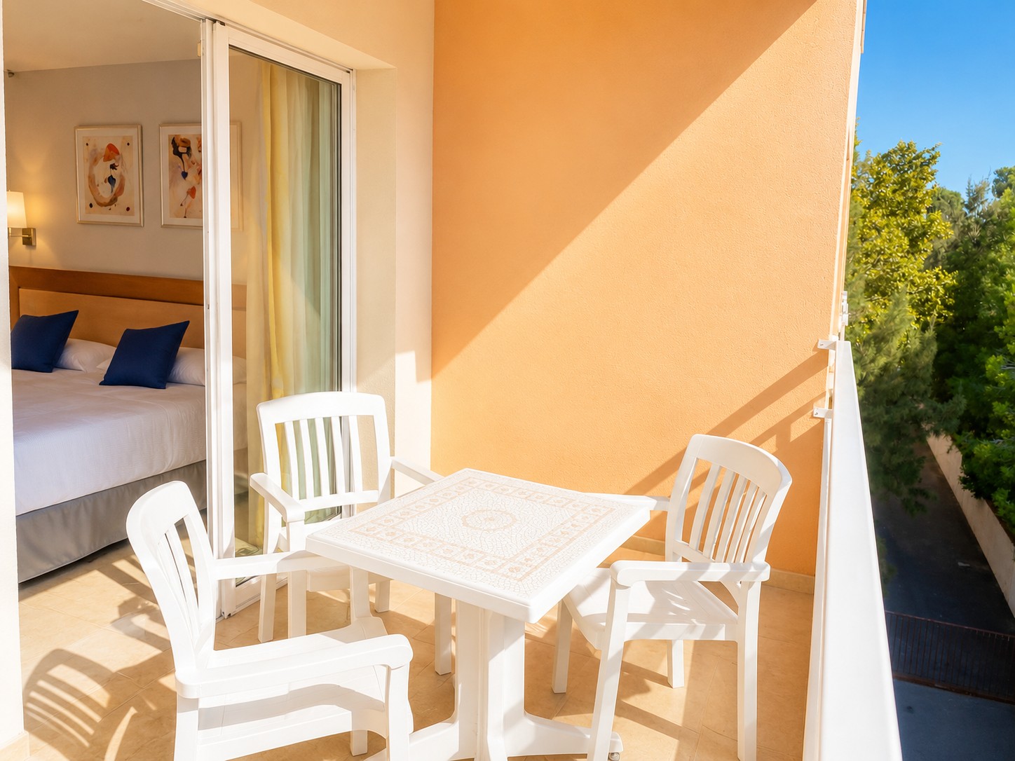 Terraza con mesa blanca y sillas, junto a una puerta con cortinas y vistas a vegetación.