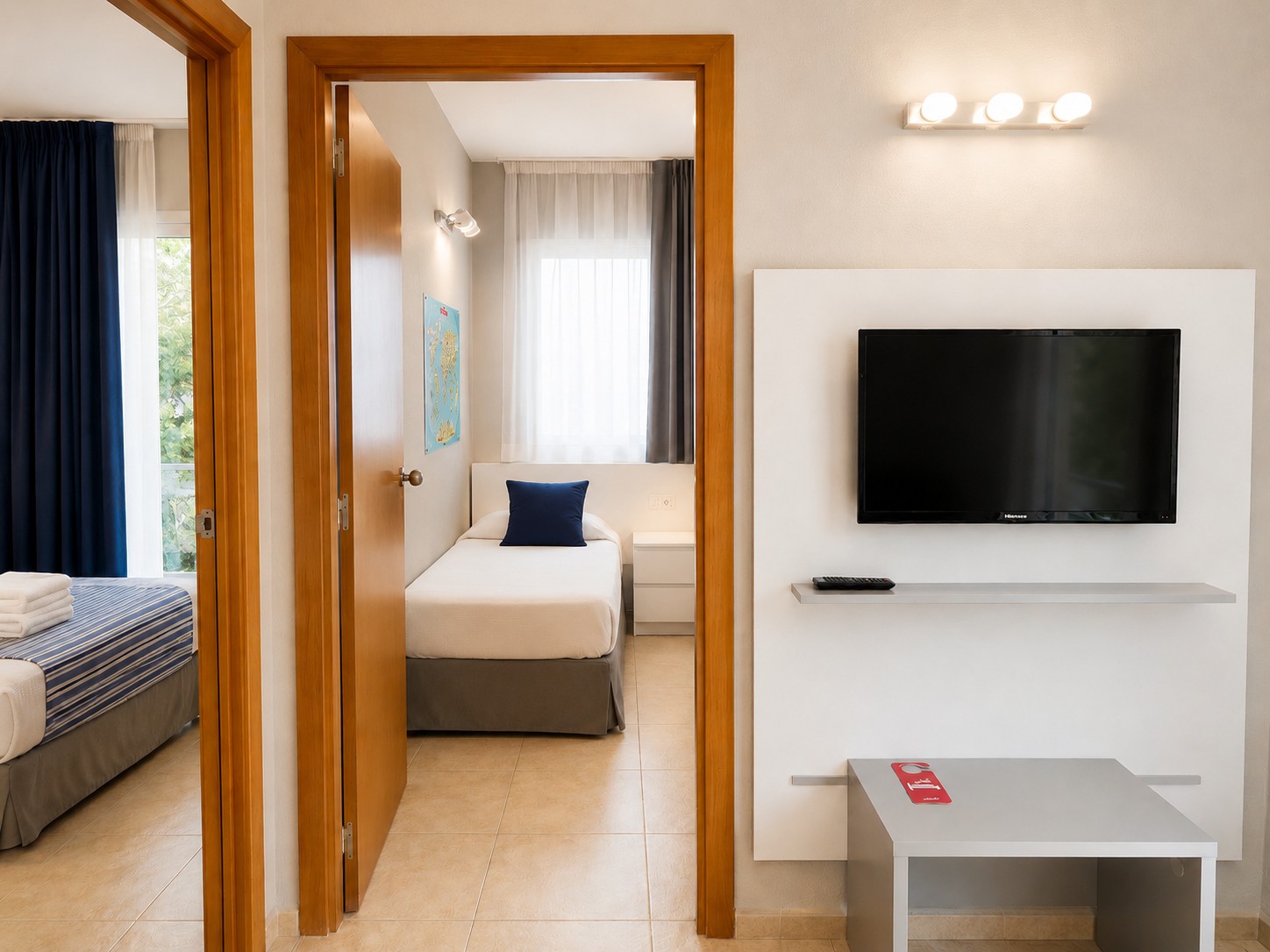 Se ve un salón con televisor de pantalla plana, mesa blanca y una puerta abierta hacia el dormitorio.