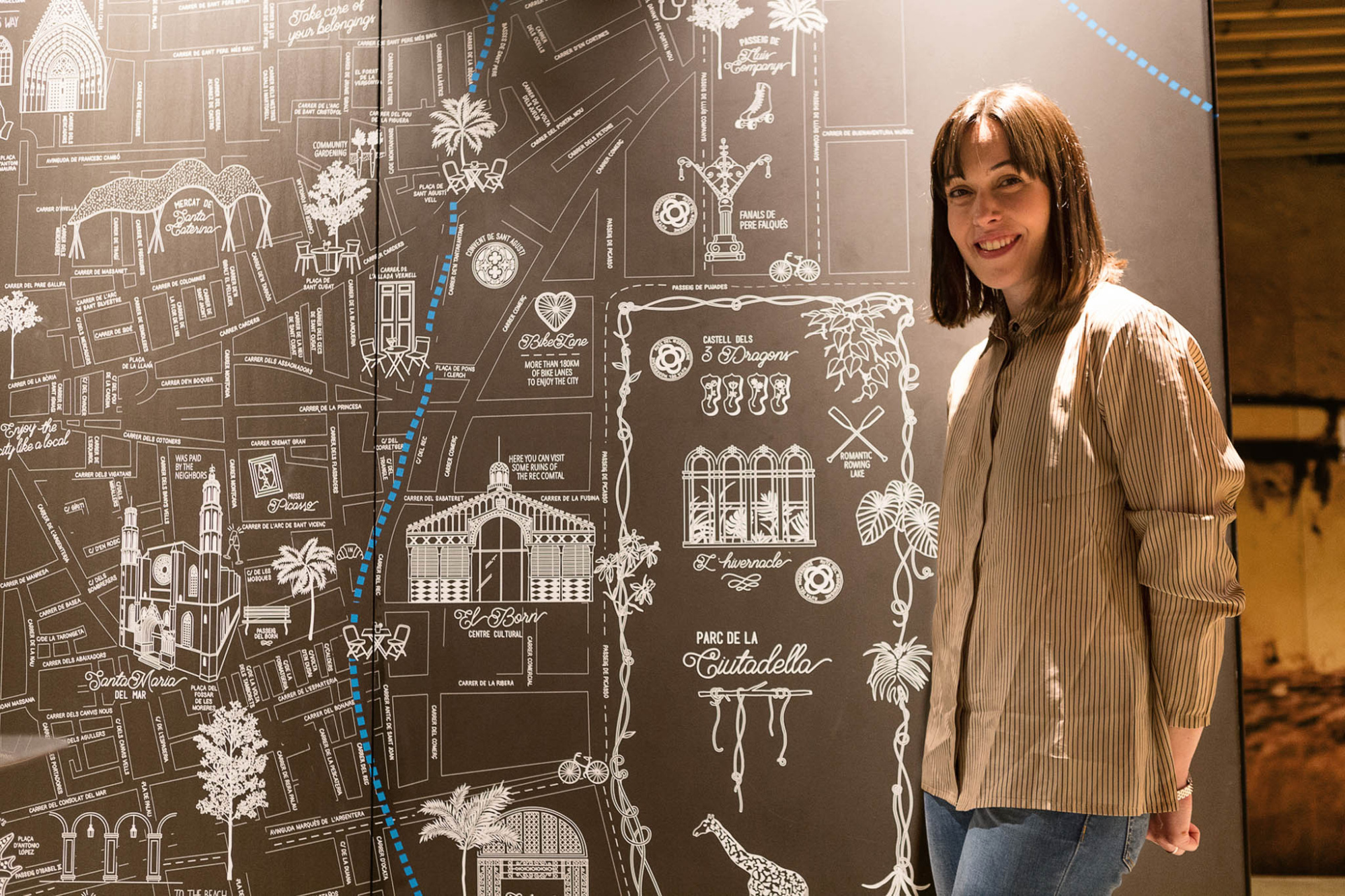 Una mujer sonríe junto a un mural con dibujos y planos en una pared oscura.