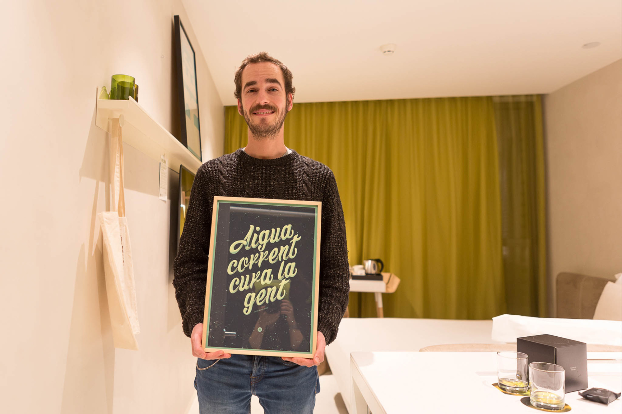 Un hombre sostiene un cuadro en una habitación con una mesa y cortinas verdes.
