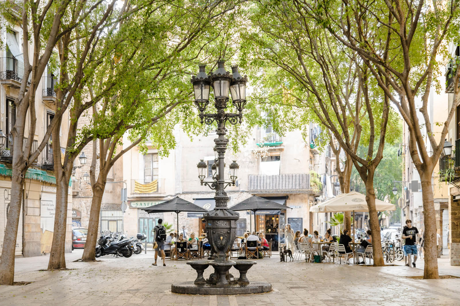 Se ve una plaza peatonal con árboles, una farola y mesas al aire libre.