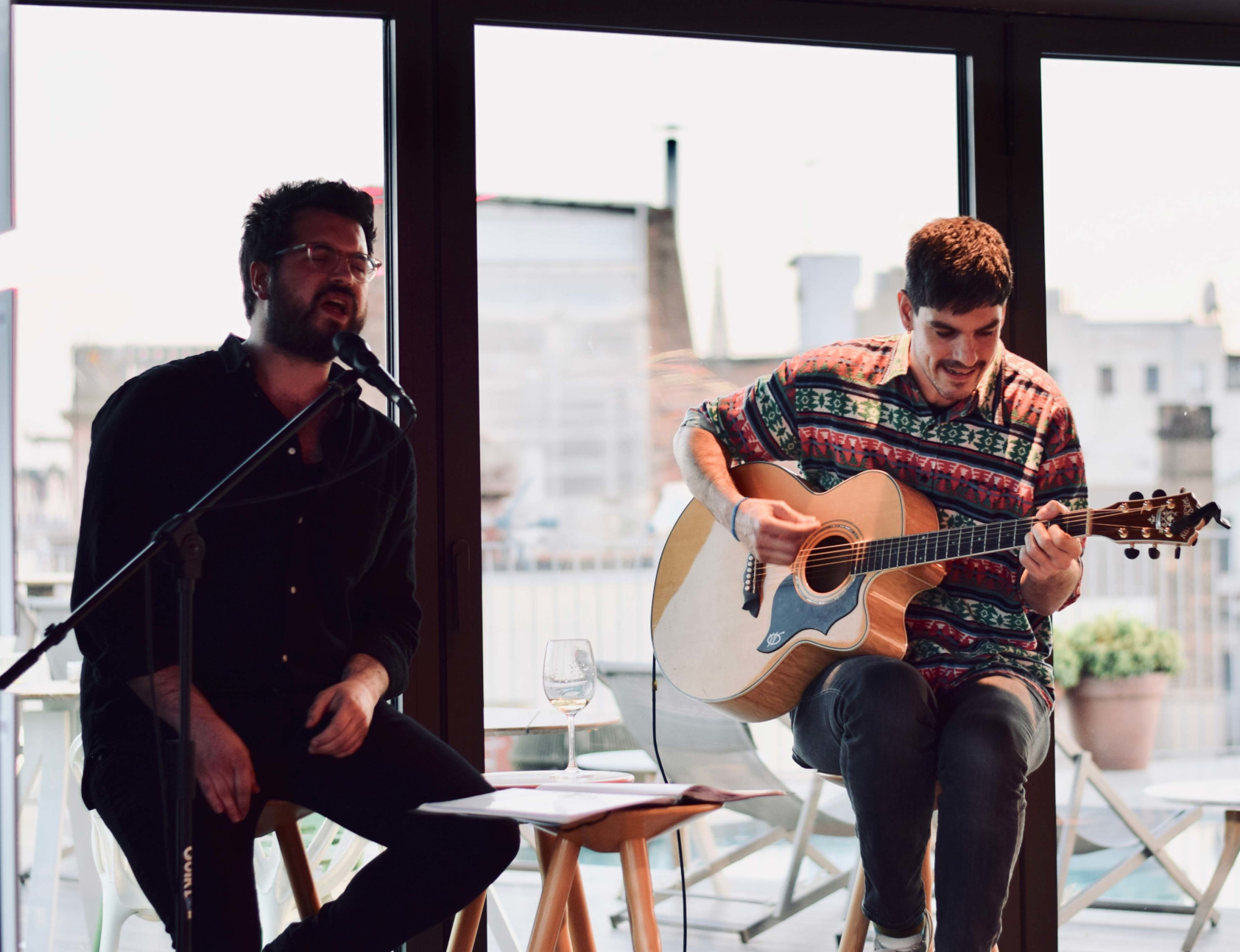 Dos músicos tocan en una terraza interior con una guitarra acústica y un micrófono.